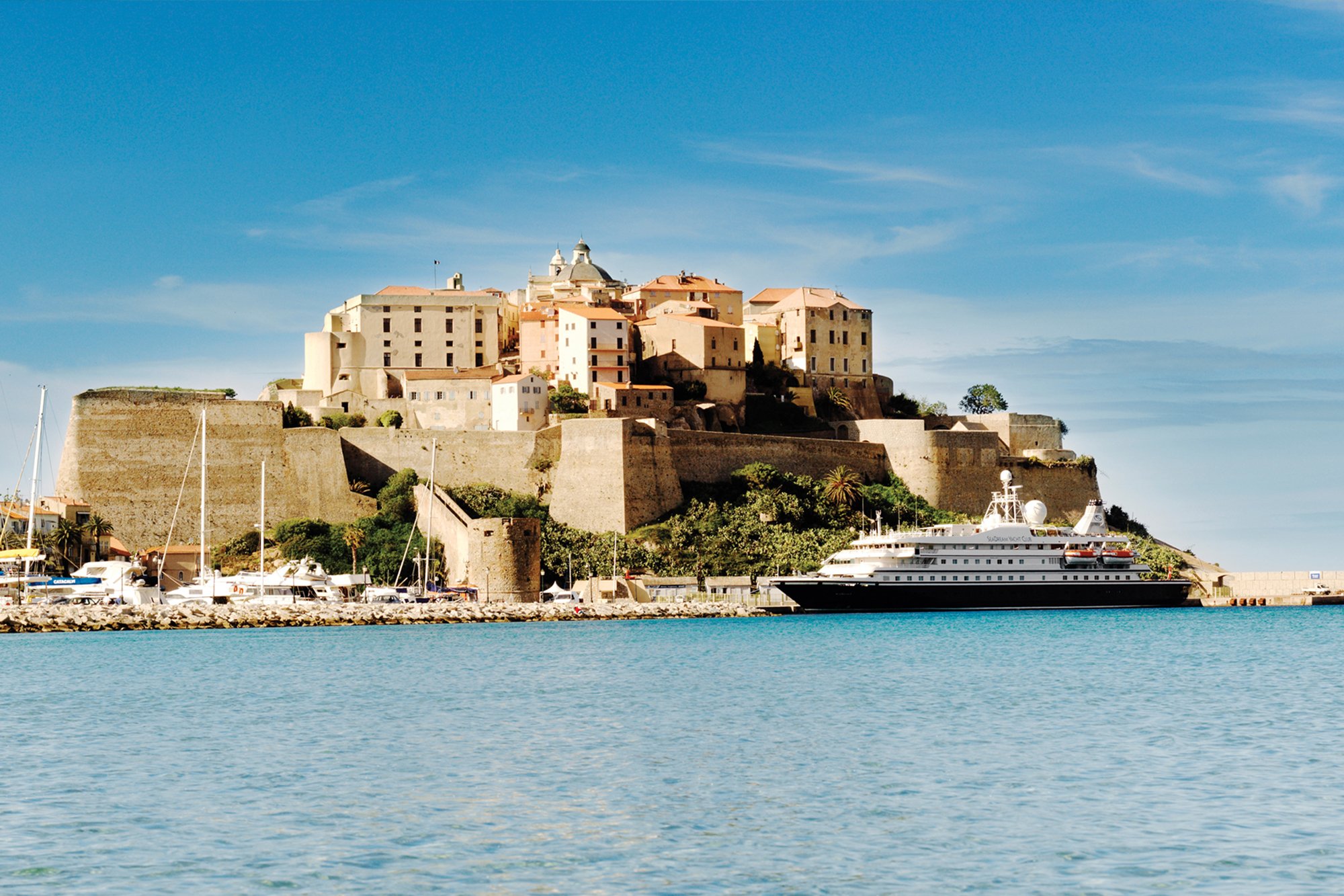 A luxury cruise ship docked at the historic port of Bonifacio, Corsica, with the impressive medieval citadel perched dramatically on the clifftop above. The fortified town features distinctive cream and terracotta architecture overlooking the turquoise Mediterranean waters, with sailboats moored in the foreground.