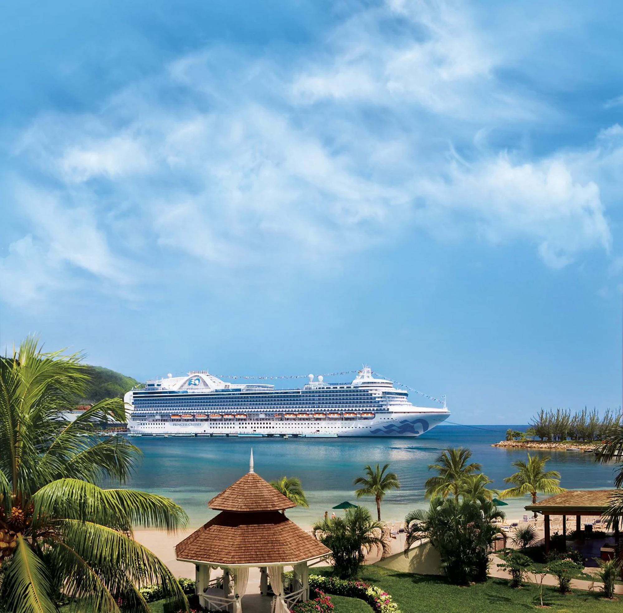 Cruise Ship at Tropical Beach Resort Port A large modern cruise ship anchors in crystal-clear turquoise waters off a pristine tropical beach resort. The scene features palm trees, thatched-roof beach pavilions, and lush landscaping in the foreground, with a bright blue sky dotted with white clouds overhead.