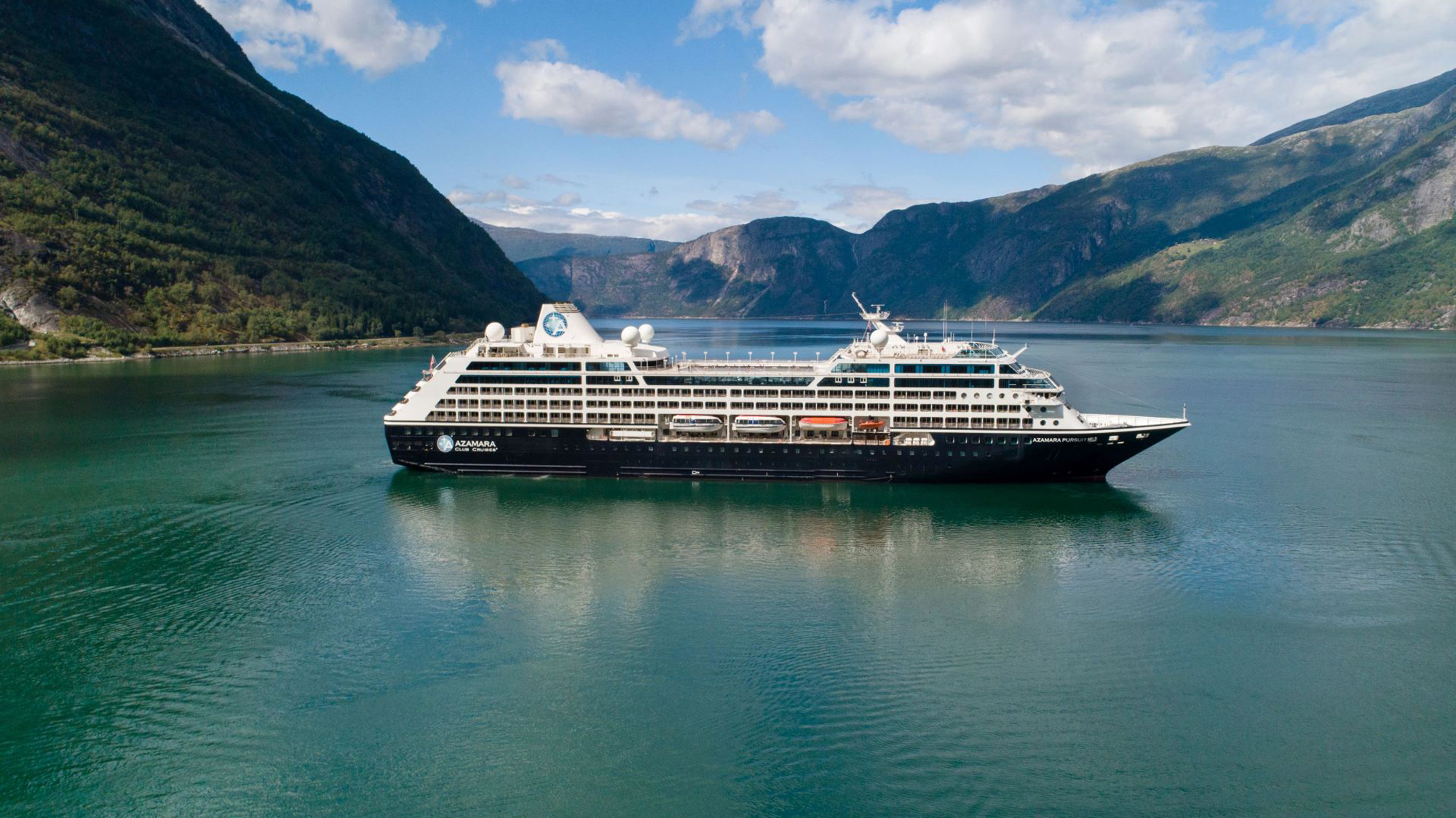 A large modern cruise ship anchors in the pristine turquoise waters of Eidfjord, surrounded by steep mountainous terrain and lush green valleys characteristic of Norwegian fjords. The dramatic landscape and clear waters showcase why this region is a premier cruise destination.