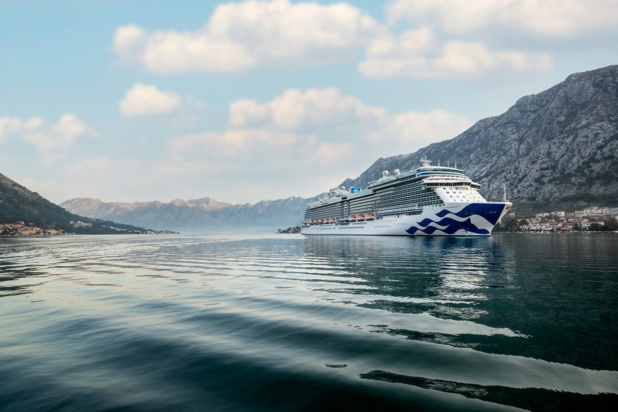 Cruise Ship in Kotor Bay, Montenegro A large modern cruise ship with distinctive blue and white livery anchors in the pristine waters of Kotor Bay, surrounded by dramatic limestone mountains and charming coastal settlements. The serene Adriatic setting showcases one of Europe's most scenic fjord-like destinations.