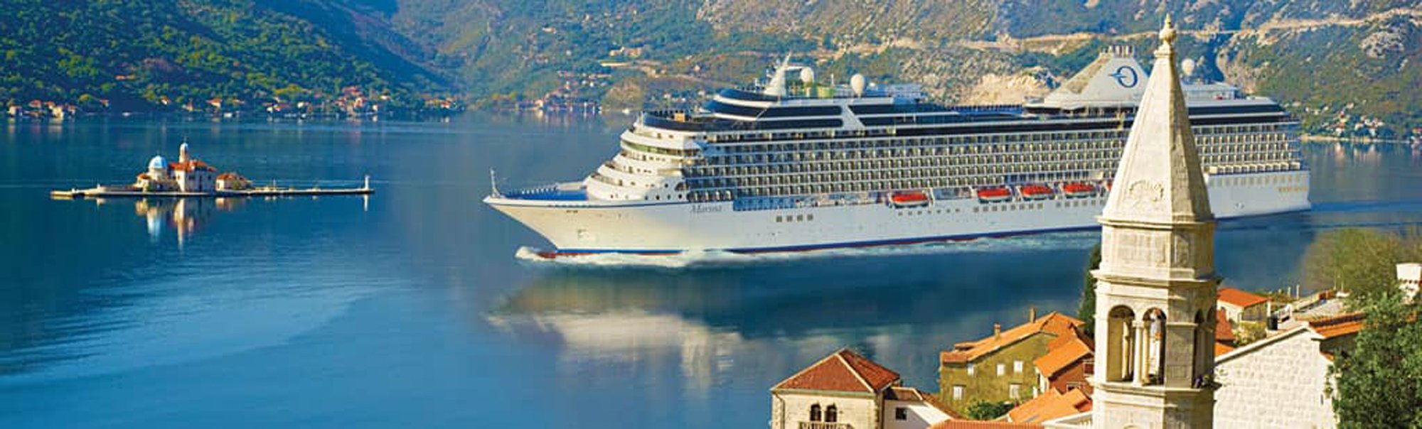A large modern cruise ship anchors in the stunning blue waters of Kotor Bay, Montenegro, with a charming coastal town featuring a bell tower in the foreground and dramatic forested mountains rising in the background. The serene alpine fjord setting showcases one of the Mediterranean's most picturesque cruise destinations.