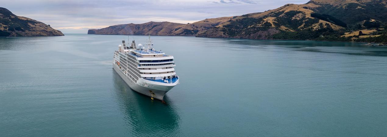 A large white cruise ship sailing through a serene blue-green water channel surrounded by rugged golden-brown hills and rocky coastline. The vessel appears to be navigating through a scenic fjord-like landscape with dramatic terrain.