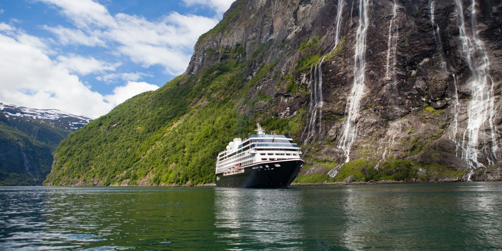 Cruise Ship in Norwegian Fjord with Waterfalls A luxury cruise ship navigates through a dramatic Norwegian fjord, surrounded by towering cliffs covered in lush green vegetation and cascading waterfalls. Snow-capped peaks are visible in the distance under a partly cloudy sky.