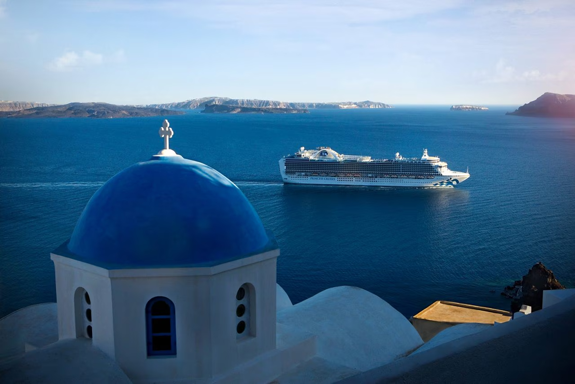 Cruise Ship in Santorini Greece A large modern cruise ship anchors in the crystalline blue waters of Santorini, Greece, viewed from the iconic whitewashed Greek Orthodox church with its distinctive blue dome perched on the clifftop. The volcanic caldera islands are visible in the distance across the brilliant Mediterranean Sea.