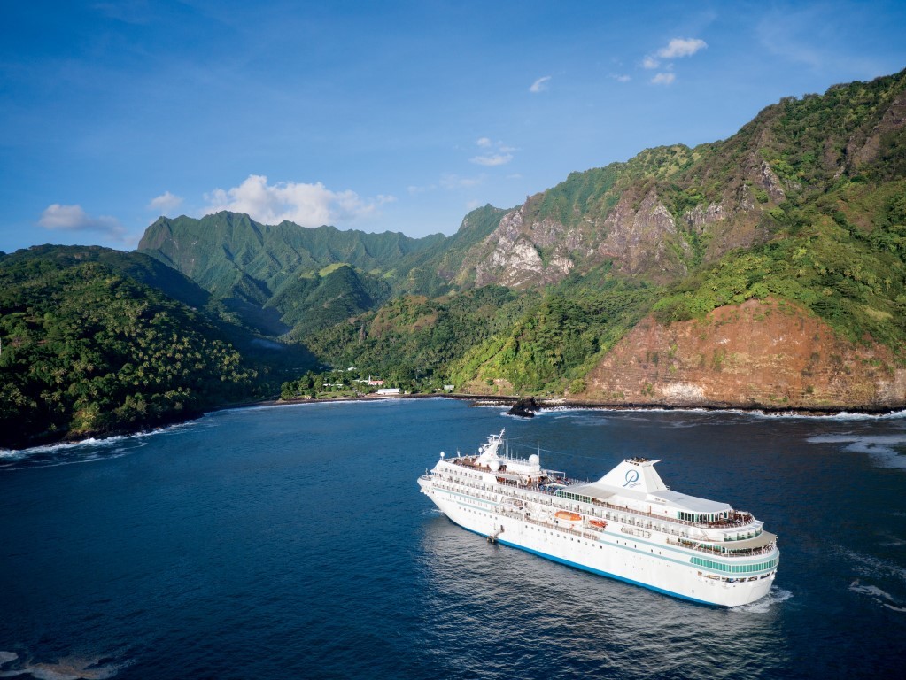 A large white cruise ship anchors in a pristine blue bay surrounded by dramatic, lush green mountains and steep volcanic cliffs in a tropical Pacific island setting. The verdant landscape features deep valleys and dense vegetation characteristic of the South Pacific region.