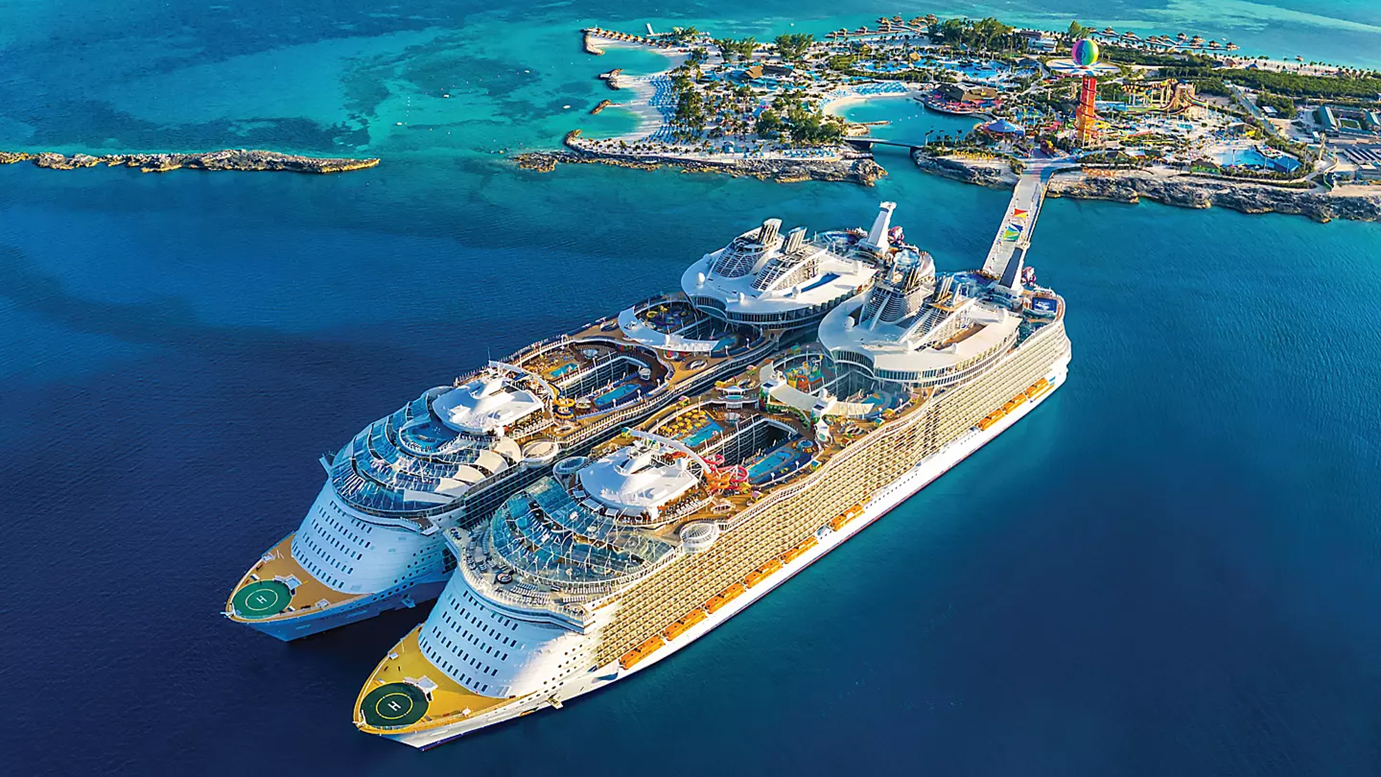 Two large modern cruise ships are anchored in crystal-clear turquoise waters off Perfect Day at CocoCay, a private island destination in the Caribbean. The island features colorful resort facilities, water parks, and recreational areas visible in the background, with the ships showcasing multiple decks, pools, and entertainment venues.