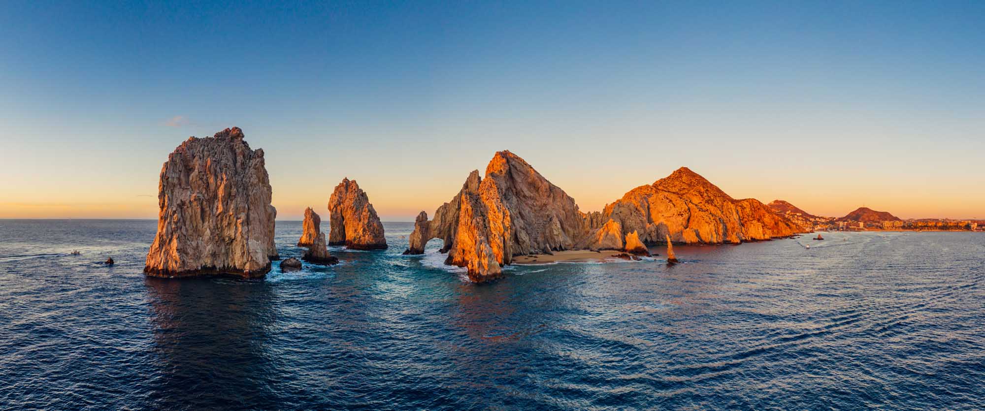 A stunning panoramic aerial view of the iconic El Arco (The Arch) rock formation at Cabo San Lucas captured during golden hour sunrise, with dramatic granite cliffs rising from turquoise Pacific waters and the coastline visible in the distance.