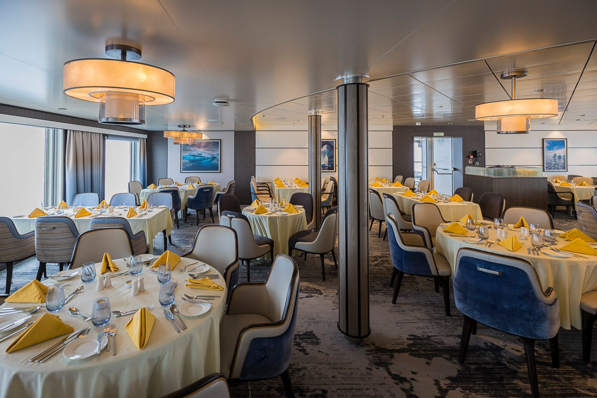 Elegant Dining Room with Gold Accents A sophisticated ship dining room featuring round tables dressed in cream linens with gold napkins, contemporary upholstered chairs in navy and beige tones, and modern pendant lighting fixtures. The space showcases floor-to-ceiling windows, decorative artwork, and sleek metal support columns, exemplifying luxury cruise ship hospitality design.