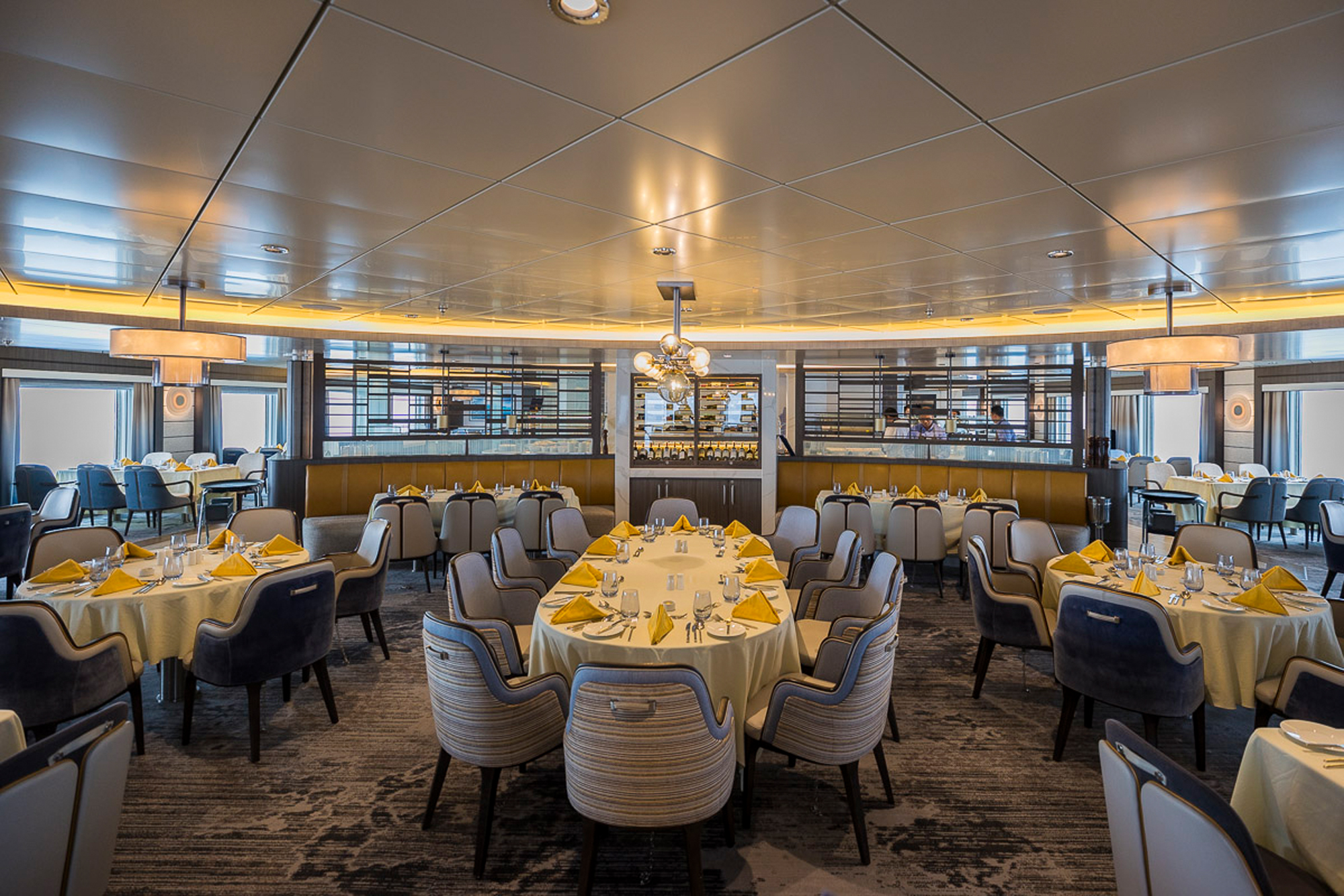 Elegant Dining Room with Ocean Views A sophisticated cruise ship dining room featuring round tables set with cream linens and golden napkins, modern striped chairs in blue and tan tones, and floor-to-ceiling windows offering panoramic ocean views. The space showcases contemporary design with a coffered ceiling, ambient lighting, and refined décor typical of luxury cruise ship dining venues.