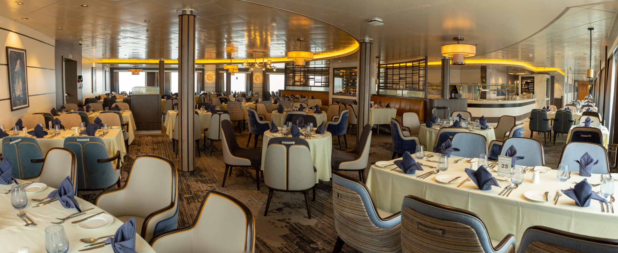 Elegant Fine Dining Restaurant Aboard Cruise Ship A sophisticated dining venue featuring multiple round tables with cream-colored linens, blue napkins, and place settings arranged throughout an expansive restaurant space. The room showcases elegant architectural elements including curved ceiling details with gold accent lighting, supporting columns, floor-to-ceiling windows with ocean views, and contemporary furnishings in neutral and blue tones, exemplifying upscale cruise ship dining.