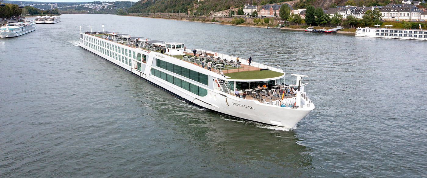 Emerald Sky River Cruise Ship in Koblenz A modern river cruise ship navigates the Rhine River in Koblenz, Germany, with multiple passenger decks, an open-air promenade, and lush greenery along the riverbanks. The vessel is surrounded by other river boats and picturesque European architecture, showcasing the charm of a traditional Rhine River cruise destination.