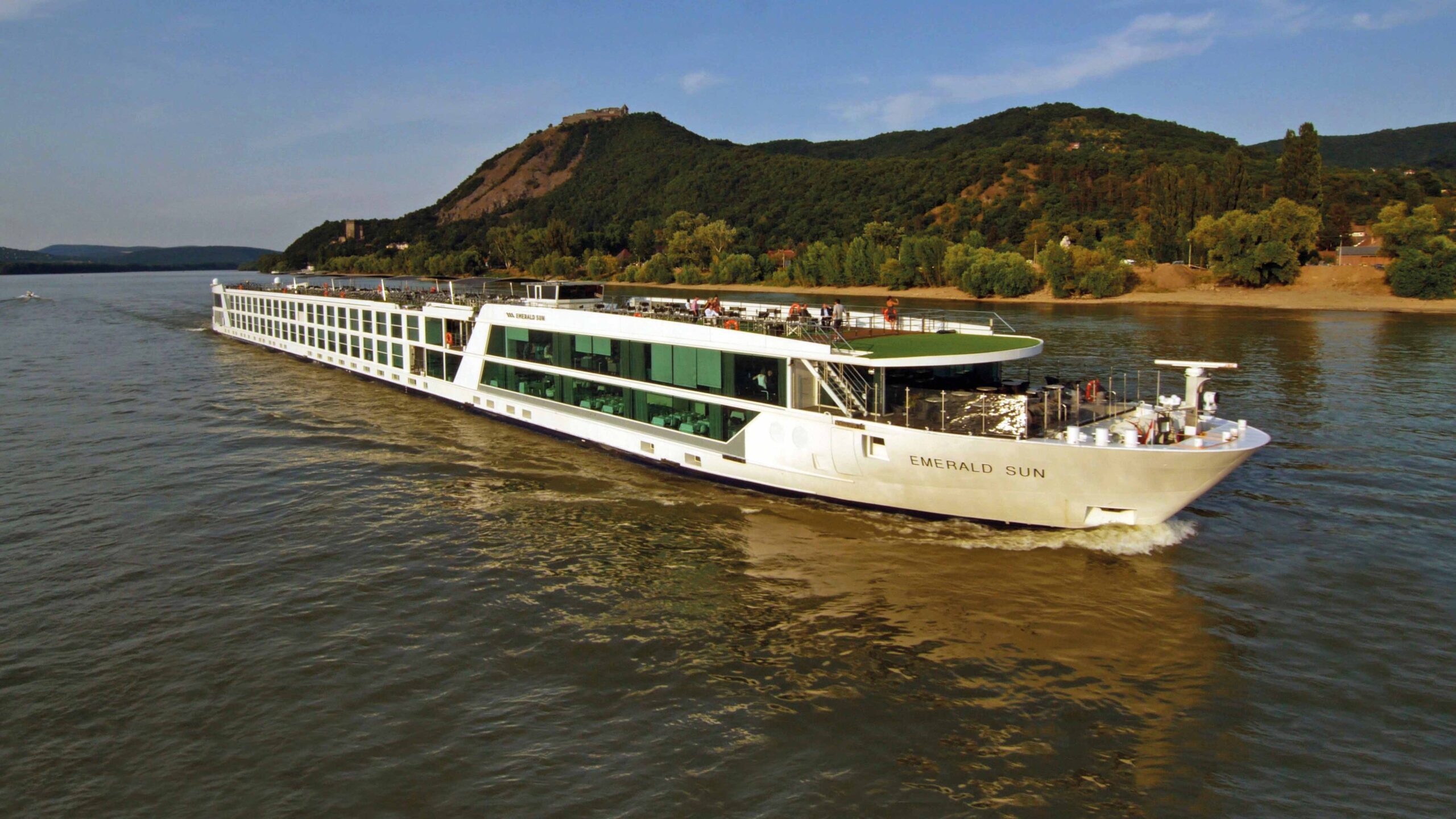 Emerald Sun River Cruise Ship on Rhine River The Emerald Sun, a modern river cruise ship with white hull and green-accented design, navigates the Rhine River with lush forested hills and a hilltop castle visible in the background. The vessel features expansive windows, an open-air sun deck, and multiple passenger amenities typical of contemporary river cruising.