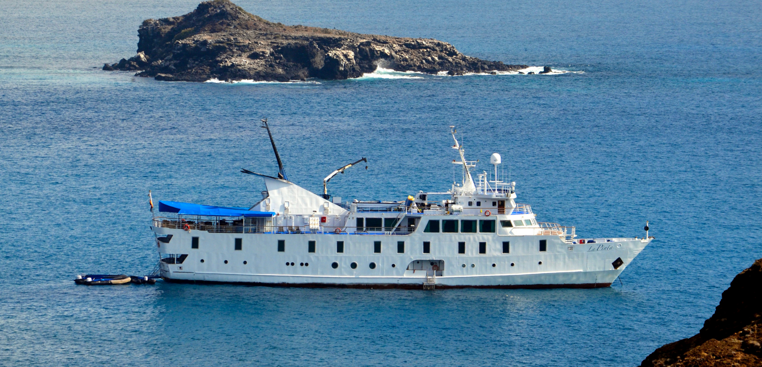 Expedition Cruise Ship Anchored Near Rocky Island A white expedition-style cruise ship is anchored in clear turquoise waters near a rocky volcanic island. The vessel appears to be a small luxury cruiser designed for expedition travel, with multiple decks and communication equipment visible, positioned in a scenic coastal setting with dramatic rocky outcrops in the background.