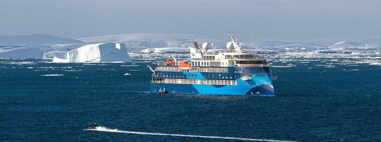 A blue and white expedition cruise ship navigates through Arctic waters alongside a massive tabular iceberg, with snow-covered Greenlandic landscape visible in the background. The vessel appears to be designed for polar exploration, featuring ice-strengthened hull and modern expedition amenities.