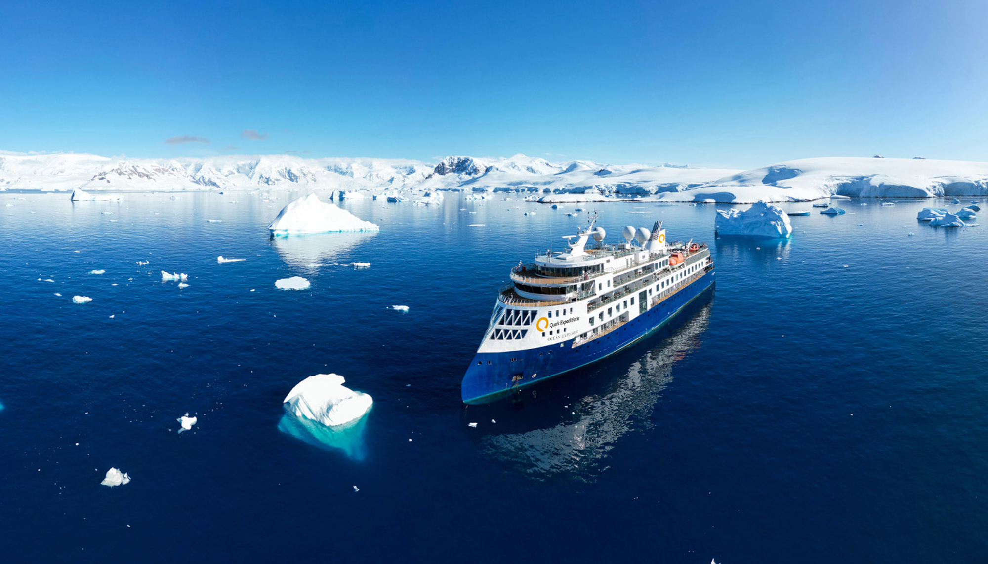 An overhead view of a polar icebreaker vessel navigating through fragmented sea ice in Arctic waters, with the ship's dark hull contrasting against white ice floes and open water channels. The image showcases the vessel's capability to break through thick ice formations in extreme polar conditions.