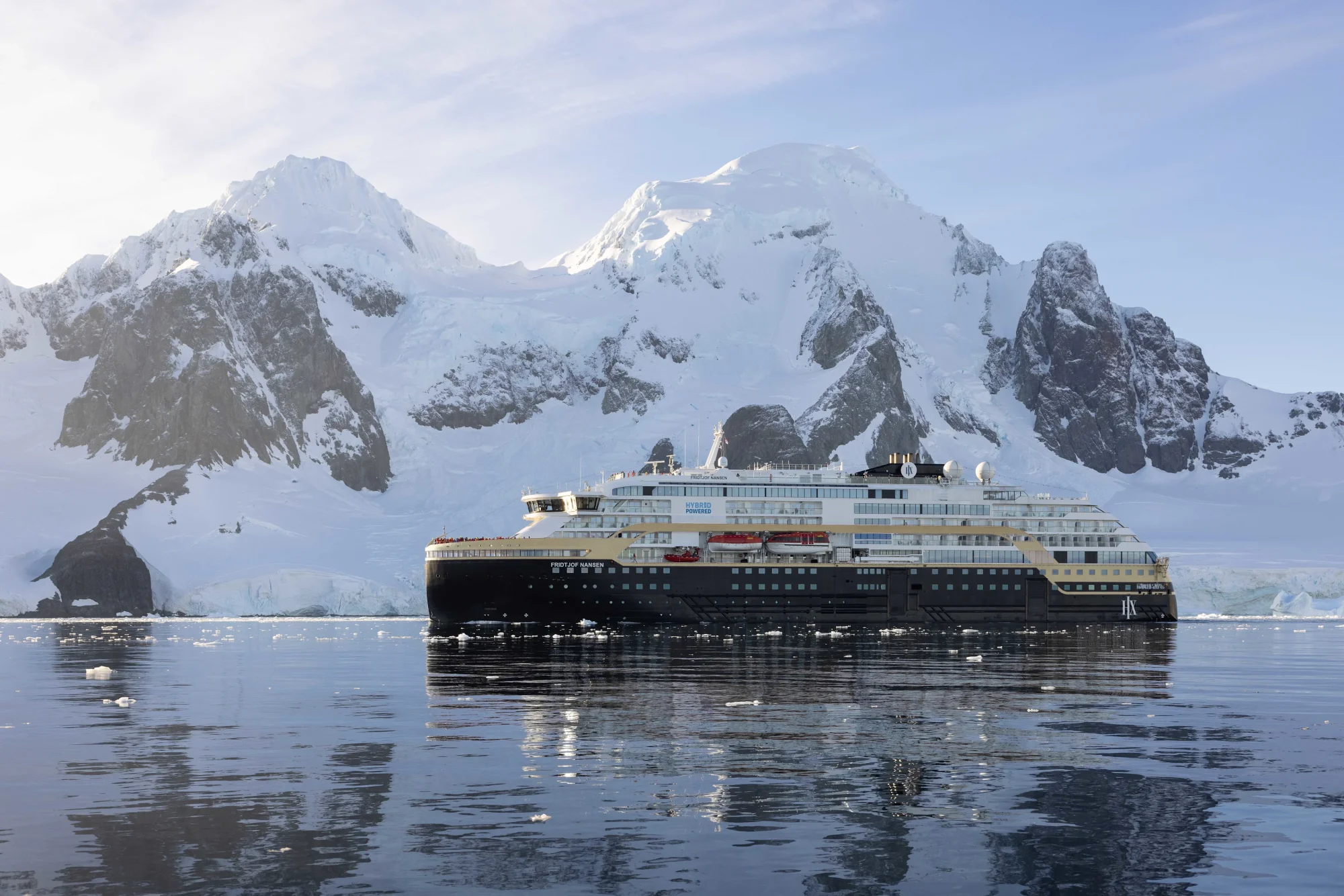 A modern expedition cruise ship navigates the pristine Antarctic waters surrounded by dramatic snow-capped peaks and rocky outcrops. The vessel is positioned against a stunning backdrop of glaciated mountains and icebergs, showcasing the remote beauty of the Antarctic Peninsula.
