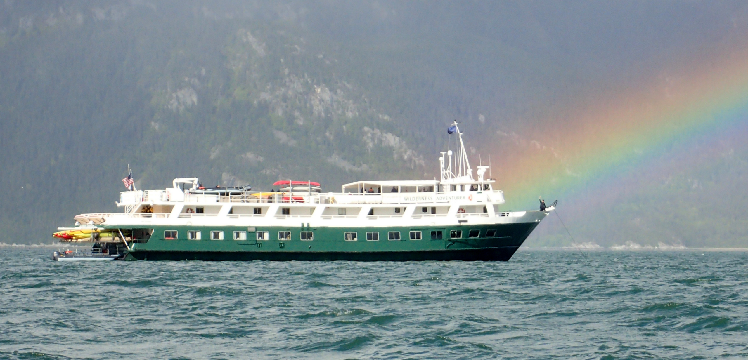 Expedition Cruise Ship in Arctic Waters A green and white expedition cruise ship navigates Arctic waters with dramatic snow-capped mountains in the background and a vibrant rainbow arcing across the misty sky. The vessel features expedition equipment and zodiac boats, typical of polar and remote destination cruising.