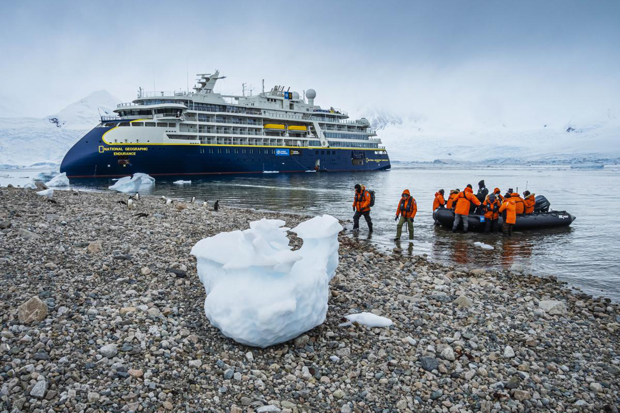 Expedition Landing at Neko Harbor, Antarctica Passengers in bright orange life jackets prepare to land via zodiac boats at a rocky beach in Neko Harbor, Antarctica, with an expedition cruise ship anchored in the background surrounded by icebergs and snow-covered mountains. The scene captures the essence of polar exploration and adventure tourism in one of Earth's most remote destinations.