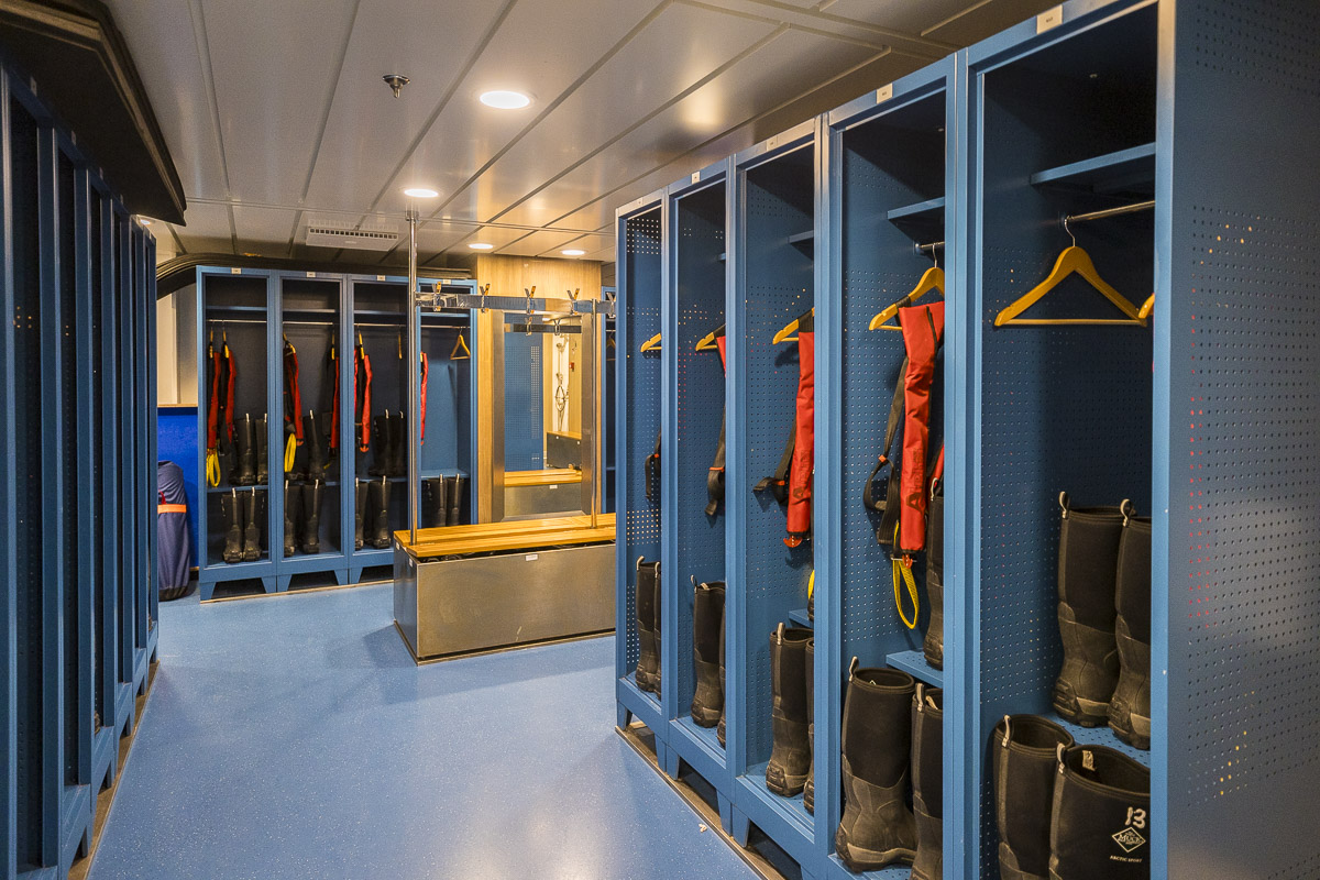 Expedition Ship Equipment Storage Room A modern mudroom and equipment storage area aboard an expedition vessel, featuring organized glass-fronted lockers with red and black cold-weather gear, wetsuits, and boots displayed on yellow hangers. The space includes a central wooden counter, blue perforated wall panels, and recessed ceiling lighting, designed for guests preparing for polar or arctic expeditions.