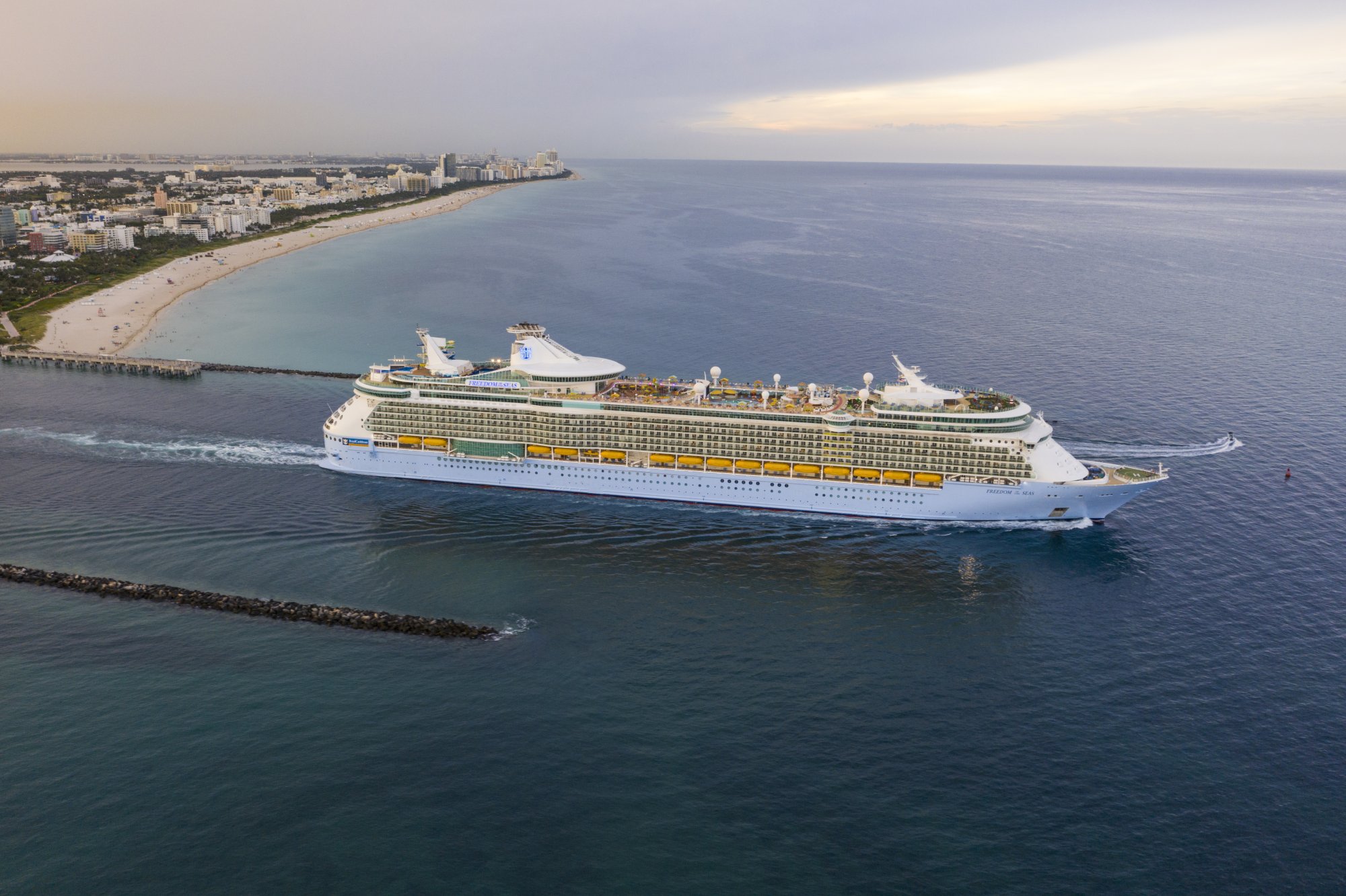 A massive Royal Caribbean cruise ship departs from the Port of Miami, with the city's beachfront skyline and sandy shores visible in the background. The white vessel with distinctive yellow and blue hull markings is captured in clear daylight, showcasing the scale and grandeur of modern cruise ship architecture.