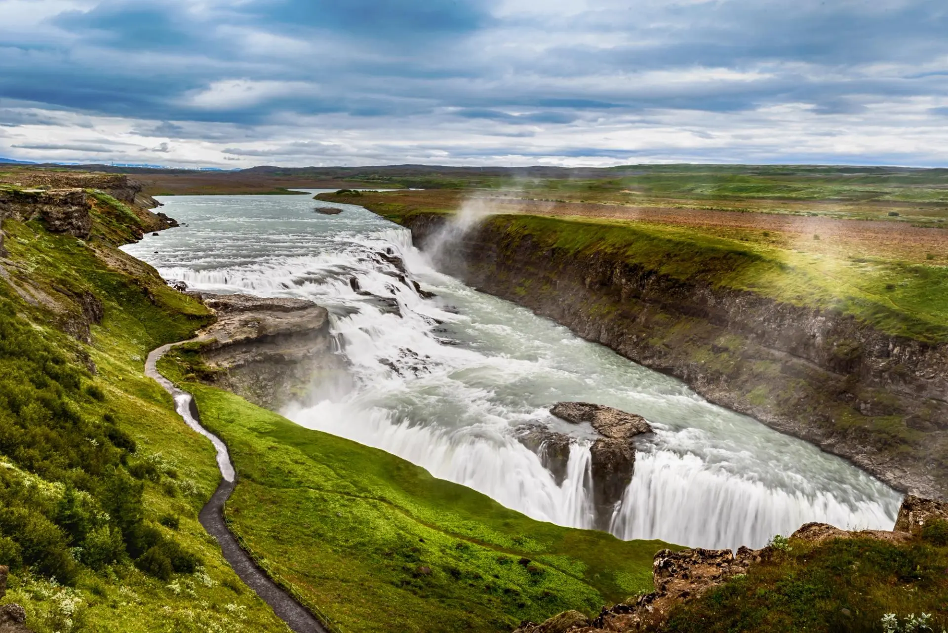 A dramatic cascading waterfall flows through a deep canyon surrounded by vibrant green moss-covered cliffs in Iceland's iconic Golden Circle region. The powerful water drops create mist clouds while winding streams cut through the lush landscape against a moody Icelandic sky.