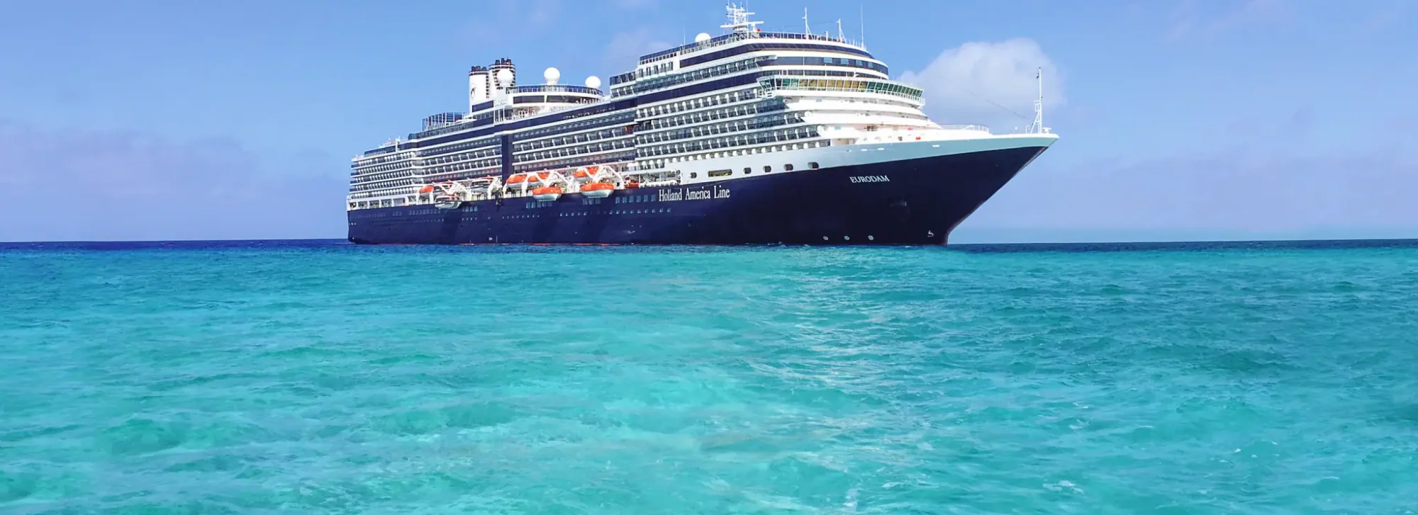 A magnificent modern cruise ship from Holland America Line anchored in crystal-clear turquoise Caribbean waters under a bright blue sky. The vessel features distinctive dark hull coloring with white superstructure and multiple decks, representing a luxury cruise experience in an idyllic tropical destination.