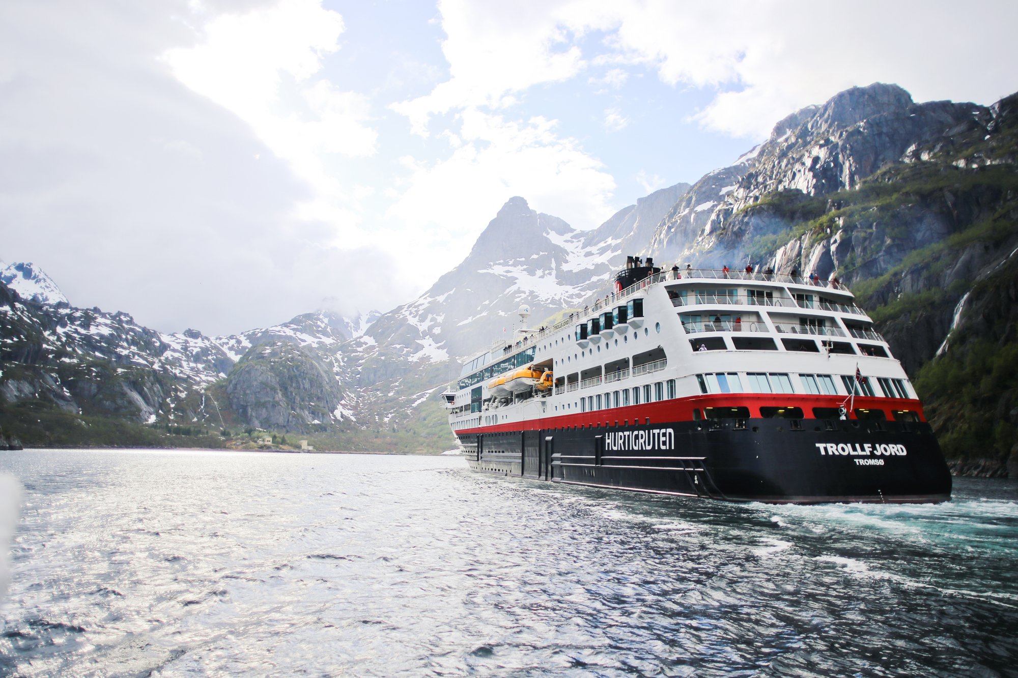The Trollfjord, a Hurtigruten cruise ship, navigates through a dramatic Norwegian fjord surrounded by snow-capped mountains, steep cliff faces, and cascading waterfalls. The modern expedition vessel with its distinctive red, white, and black hull is dwarfed by the towering landscape, showcasing the spectacular scenery of Norway's fjord region.