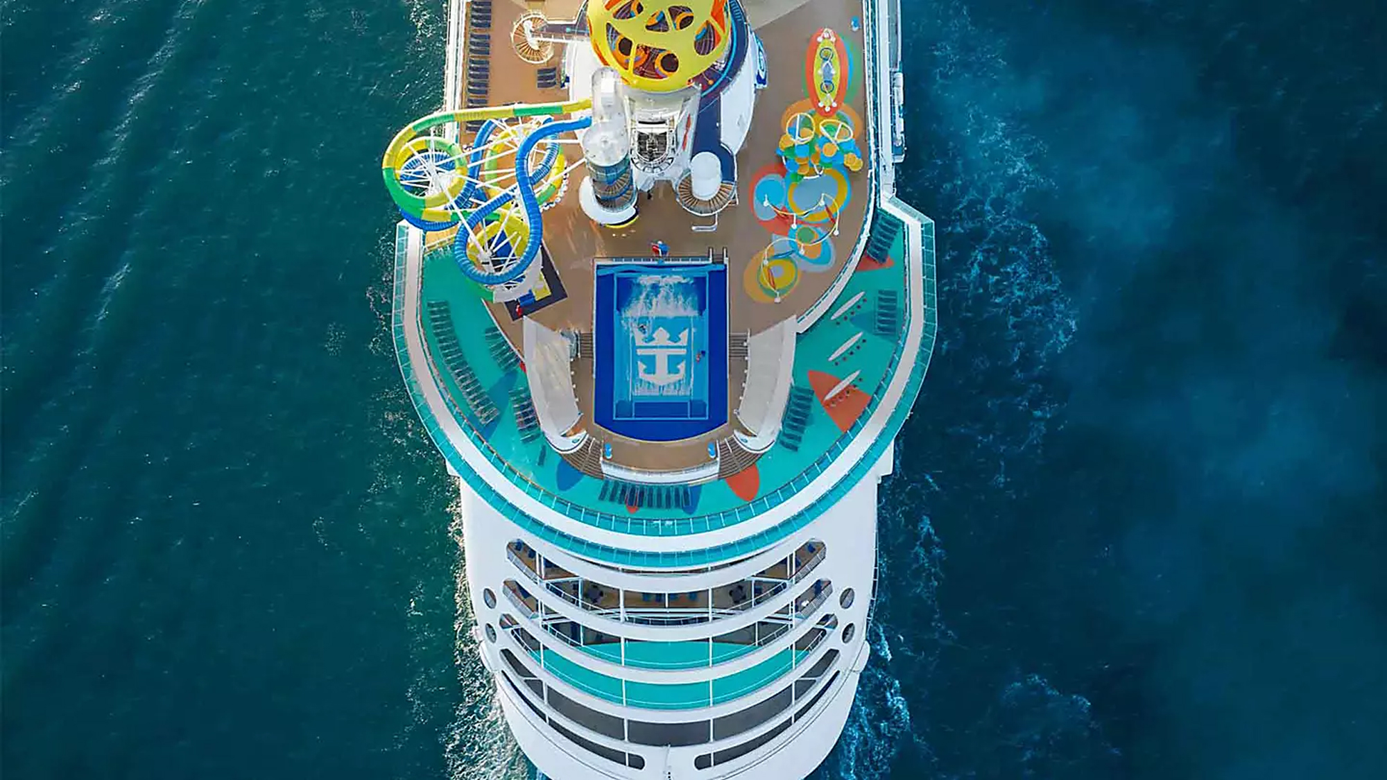 An overhead aerial view of a large cruise ship's upper deck featuring vibrant water slides, recreational pools, and colorful play structures. The turquoise and white vessel is surrounded by deep blue ocean water, showcasing the extensive entertainment facilities and deck amenities typical of modern cruise ship design.