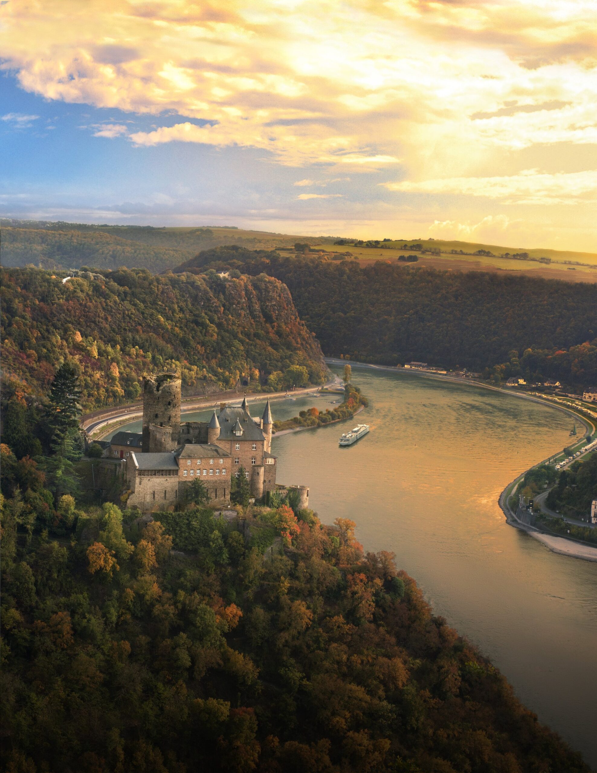 A stunning aerial view of Katz Castle perched dramatically on a forested cliff overlooking the Middle Rhine River Valley in Germany. A Viking Longship cruise vessel navigates the golden waters below, surrounded by autumn-colored forests and rolling hills, with the castle's distinctive towers silhouetted against a warm sunset sky.