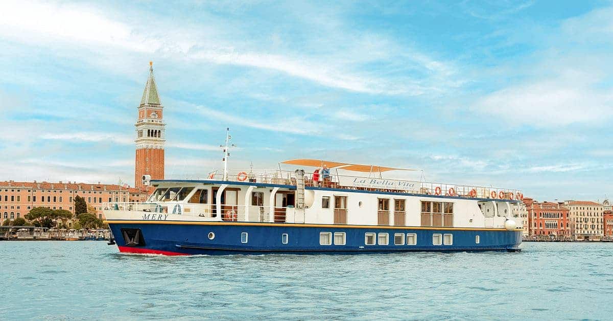 A modern river cruise vessel named 'La Bella Vita' navigates the turquoise waters of Venice's Grand Canal, with the iconic St. Mark's Basilica campanile (bell tower) and historic Venetian architecture visible in the background under a bright blue sky.