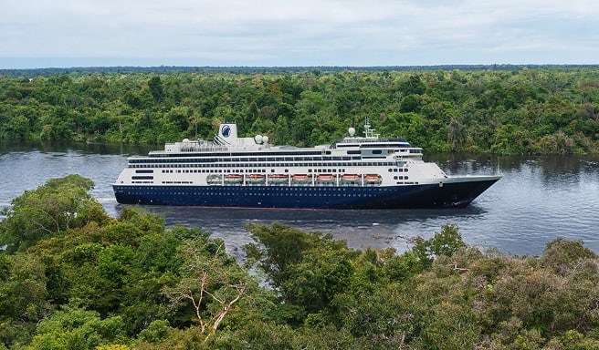 A modern ocean-going cruise ship navigates a wide river surrounded by lush tropical rainforest canopy. The vessel features a white superstructure with dark hull, photographed from an elevated vantage point with dense green vegetation framing the scene.