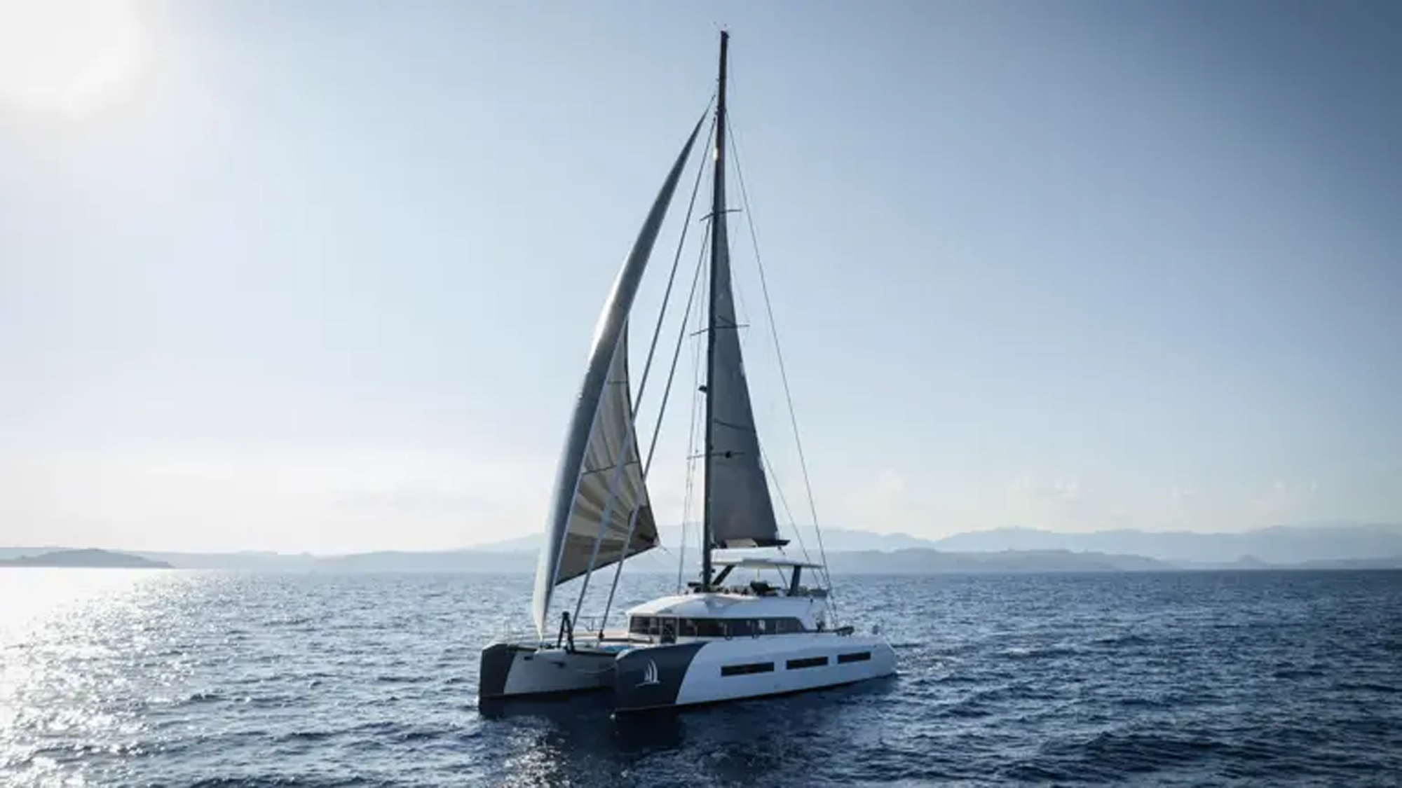 A modern luxury catamaran with white hull and black accents sails under full canvas in clear Mediterranean waters, with mountainous coastline visible in the distance under blue skies. The vessel features contemporary design with multiple deck levels and professional sailing rigging.