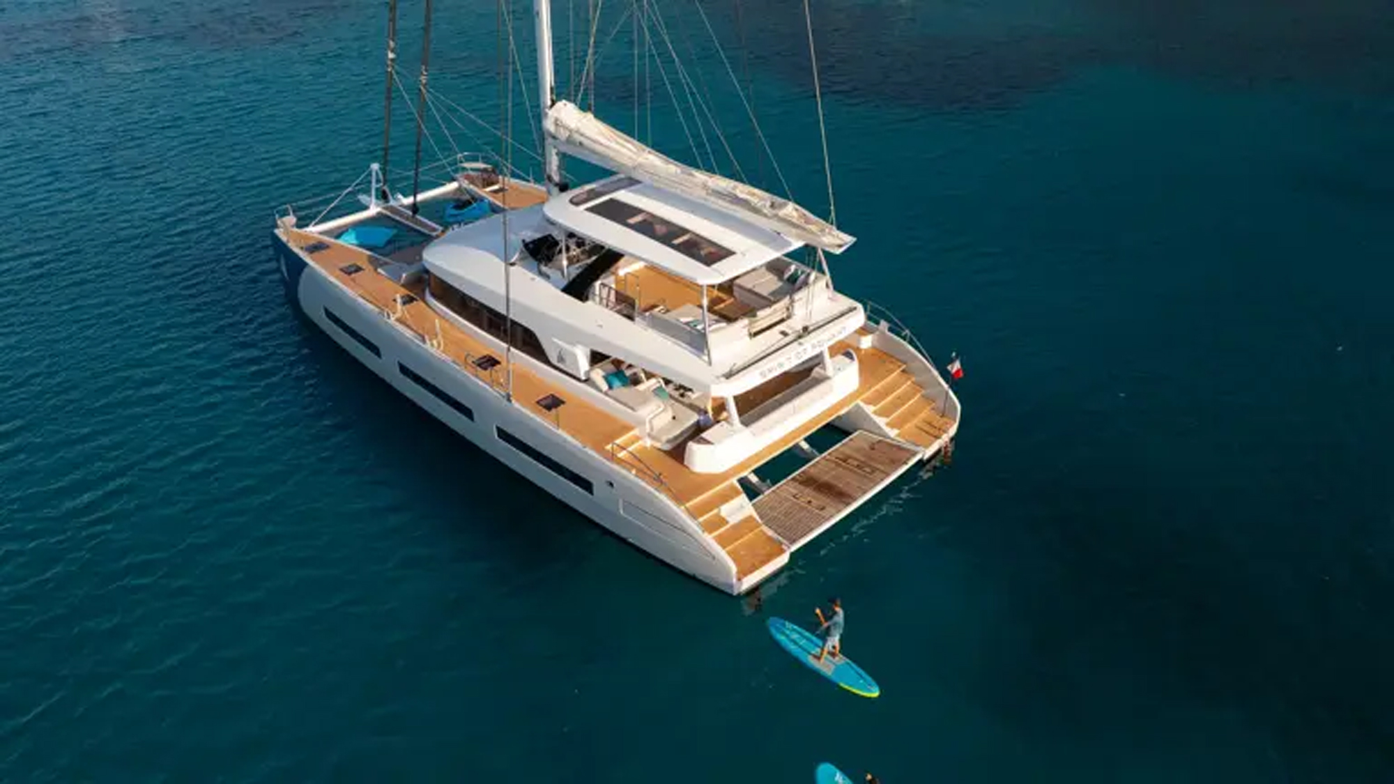 A modern white and natural wood luxury catamaran yacht anchored in stunning turquoise waters, with passengers enjoying water activities including paddleboarding and swimming in the crystal-clear Mediterranean or tropical sea.