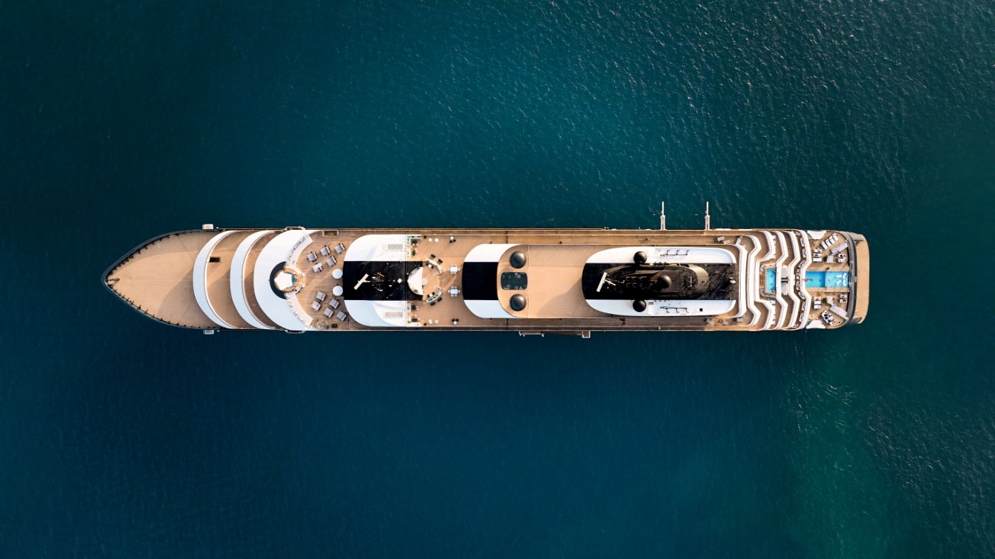 An overhead aerial view of a large luxury cruise ship on turquoise water, showcasing the full length of the vessel with its distinctive deck layout, multiple pools, and white superstructure against the deep teal ocean.
