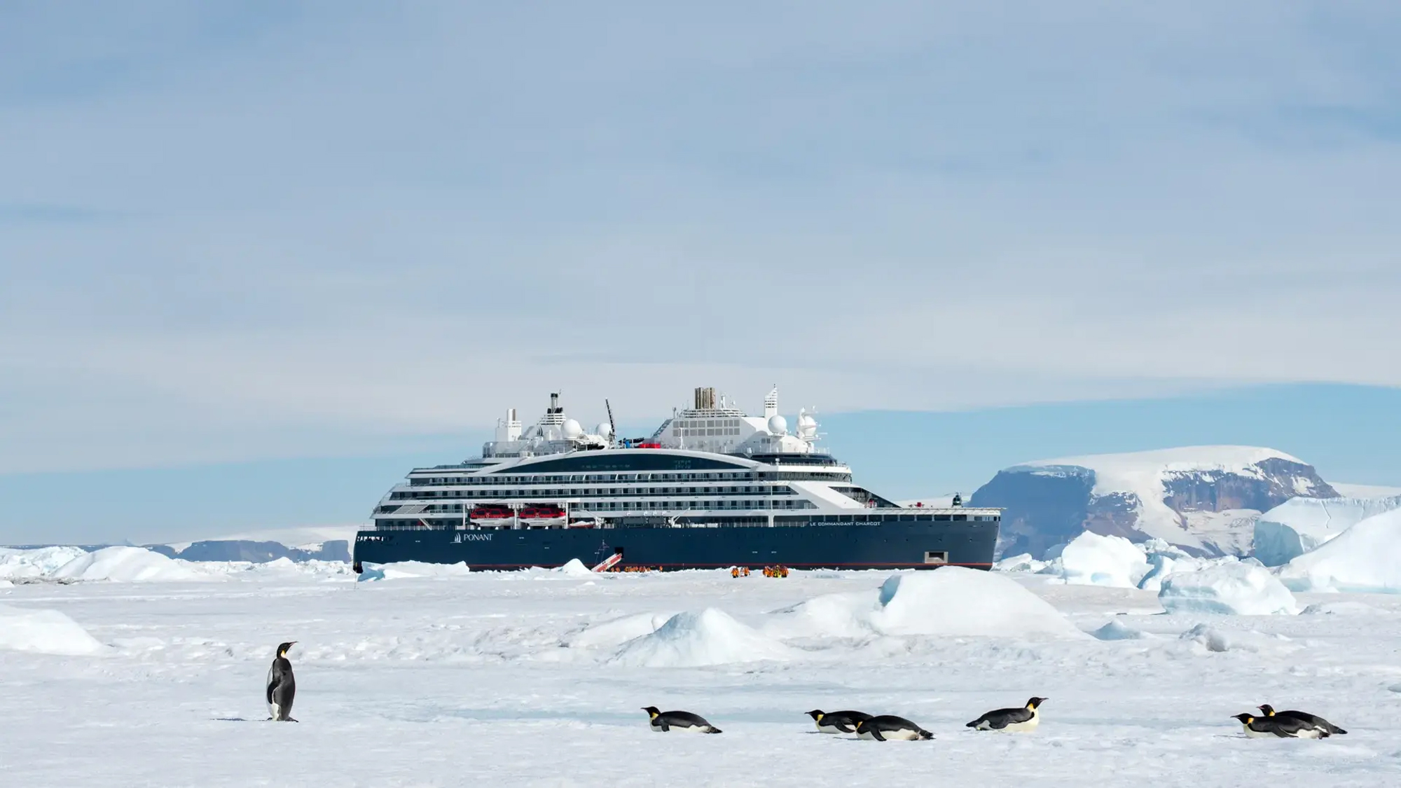 A modern expedition cruise ship navigates through the icy waters of the Weddell Sea in Antarctica, surrounded by massive white icebergs and pack ice. Adelie penguins are visible on the frozen surface in the foreground, highlighting the pristine wildlife and extreme polar environment of Snow Hill Island.
