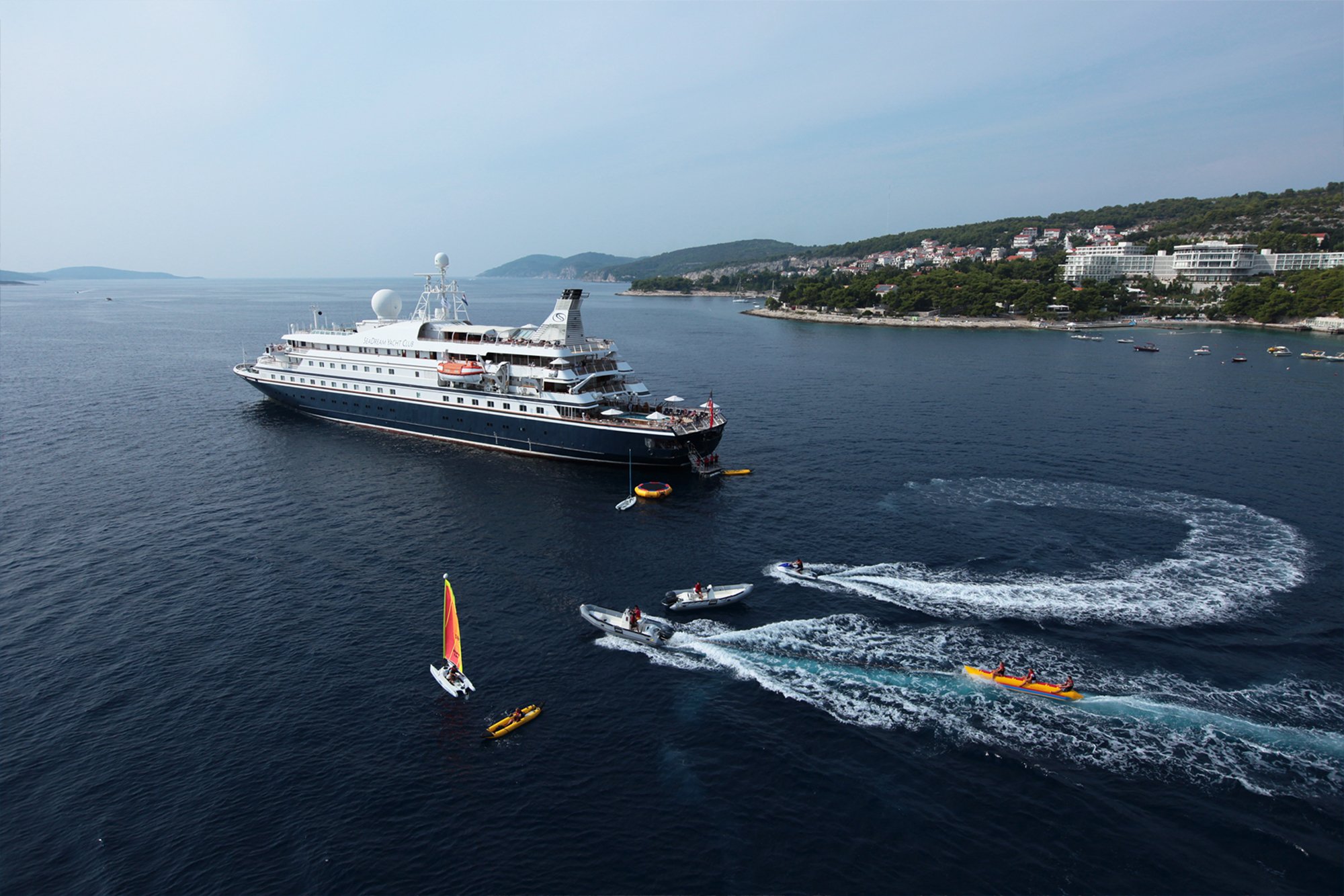 A large expedition cruise ship with distinctive dark hull and white superstructure is anchored in crystal-clear turquoise waters off a scenic Mediterranean coastal town. Small water sports boats and kayaks are visible in the foreground, while a picturesque hillside village with white buildings and lush green vegetation rises behind the vessel.
