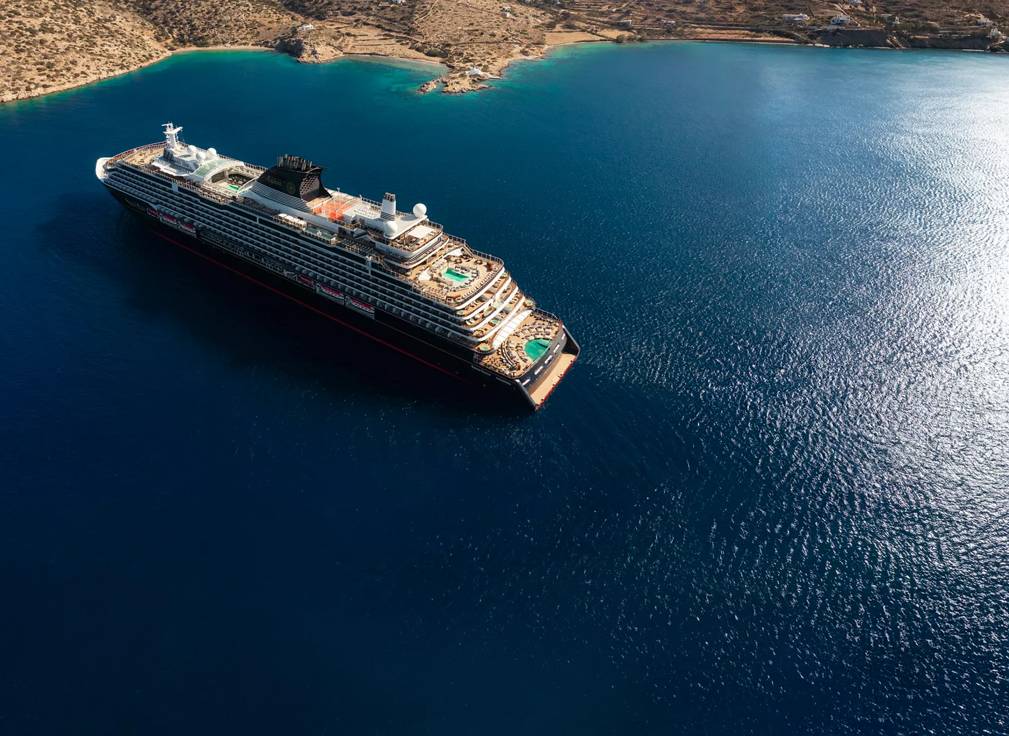 Luxury Cruise Ship Anchored in Mediterranean Waters An aerial view of a large modern cruise ship anchored in crystal-clear turquoise Mediterranean waters near an arid, rocky coastline. The ship features multiple deck levels with pools, hot tubs, and recreational areas visible from above, showcasing the expansive leisure amenities of contemporary cruise vessel design.