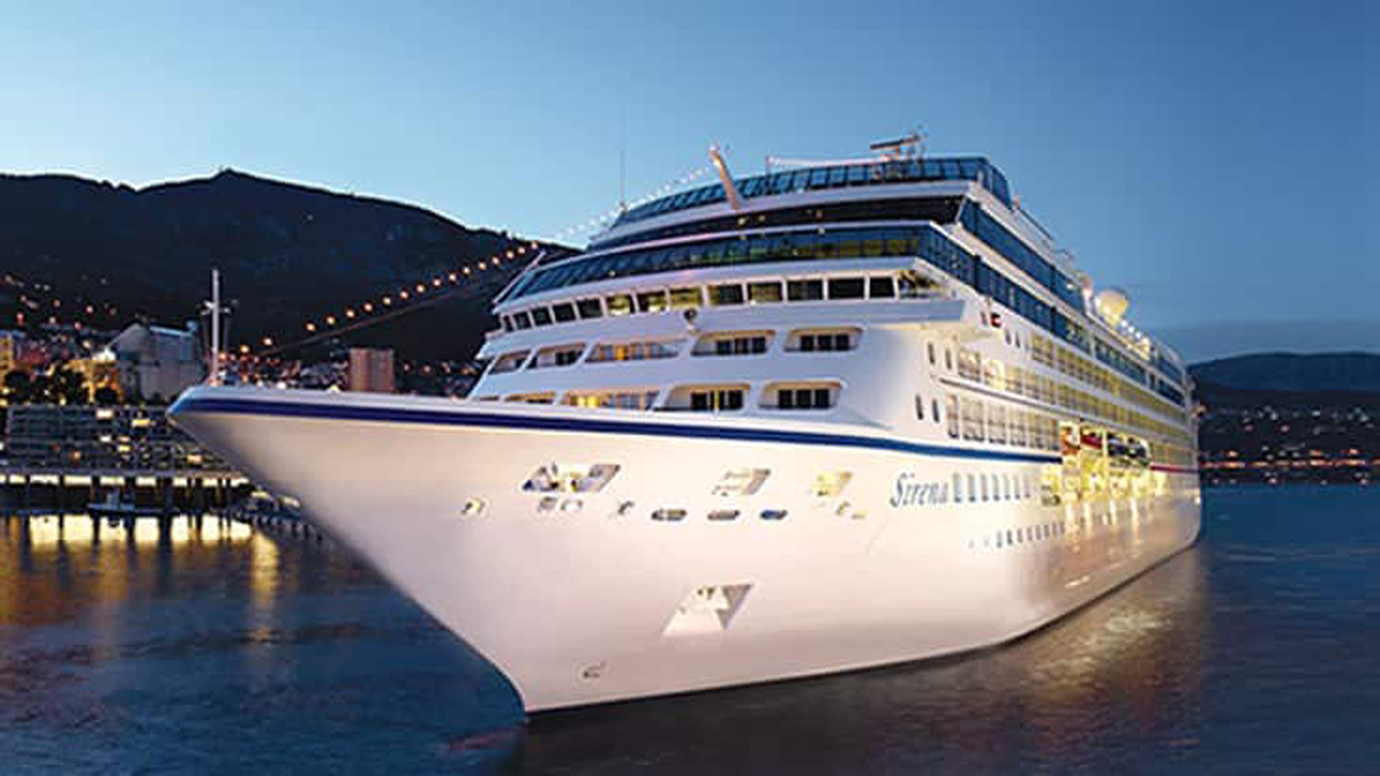 A large, modern luxury cruise ship with distinctive white hull and blue upper decks is moored at a picturesque port nestled against snow-capped mountains during twilight hours. The vessel's illuminated windows and the harbor's evening lights create a serene scene, with mountainous terrain and waterfront buildings visible in the background.