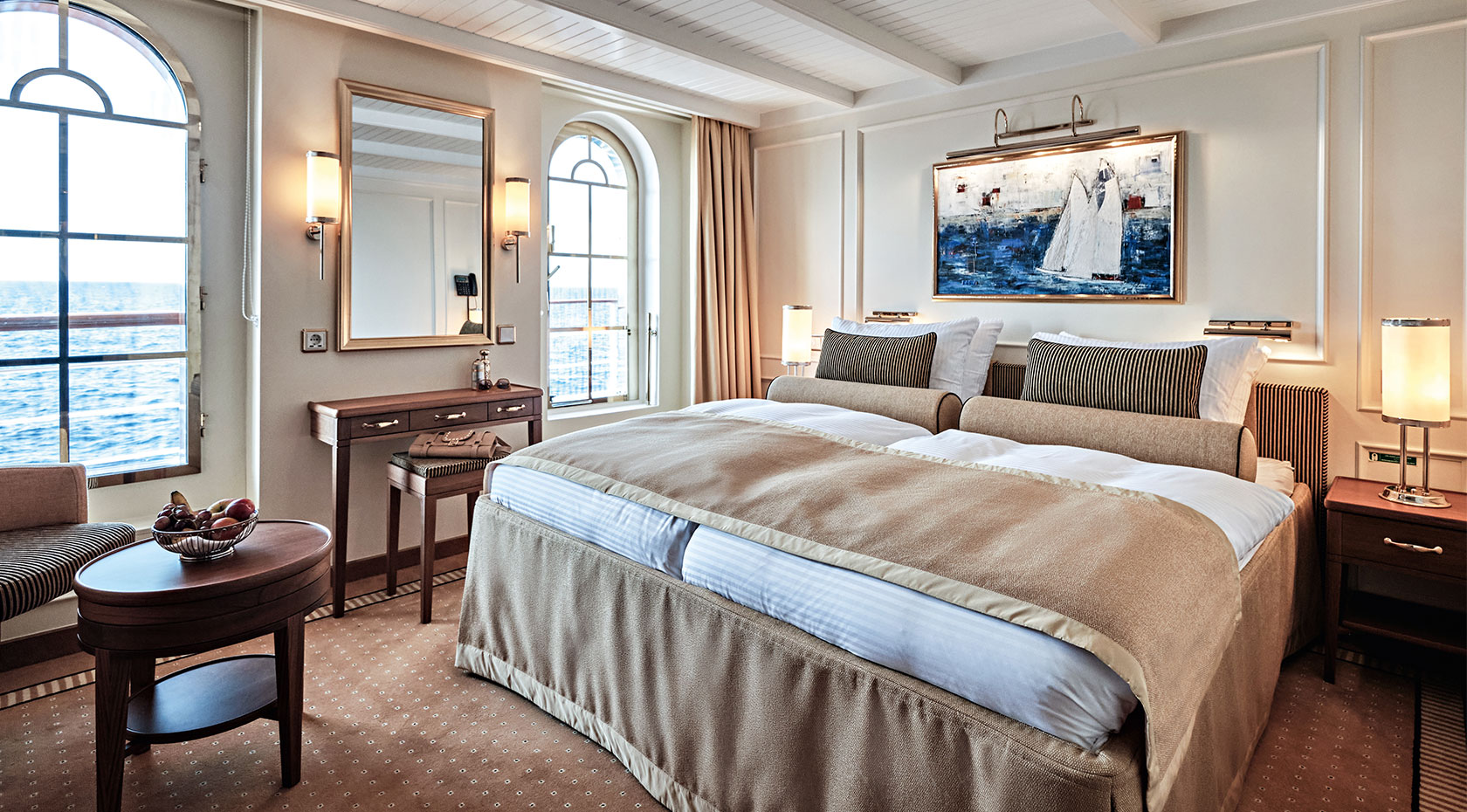 Luxury Cruise Ship Ocean View Stateroom An elegantly appointed oceanview stateroom aboard a luxury cruise ship, featuring a premium bed with neutral bedding, floor-to-ceiling windows overlooking the sea, a seating area with striped upholstered furniture, and nautical-themed décor including a sailboat artwork. The sophisticated cabin showcases modern amenities and refined coastal design elements.