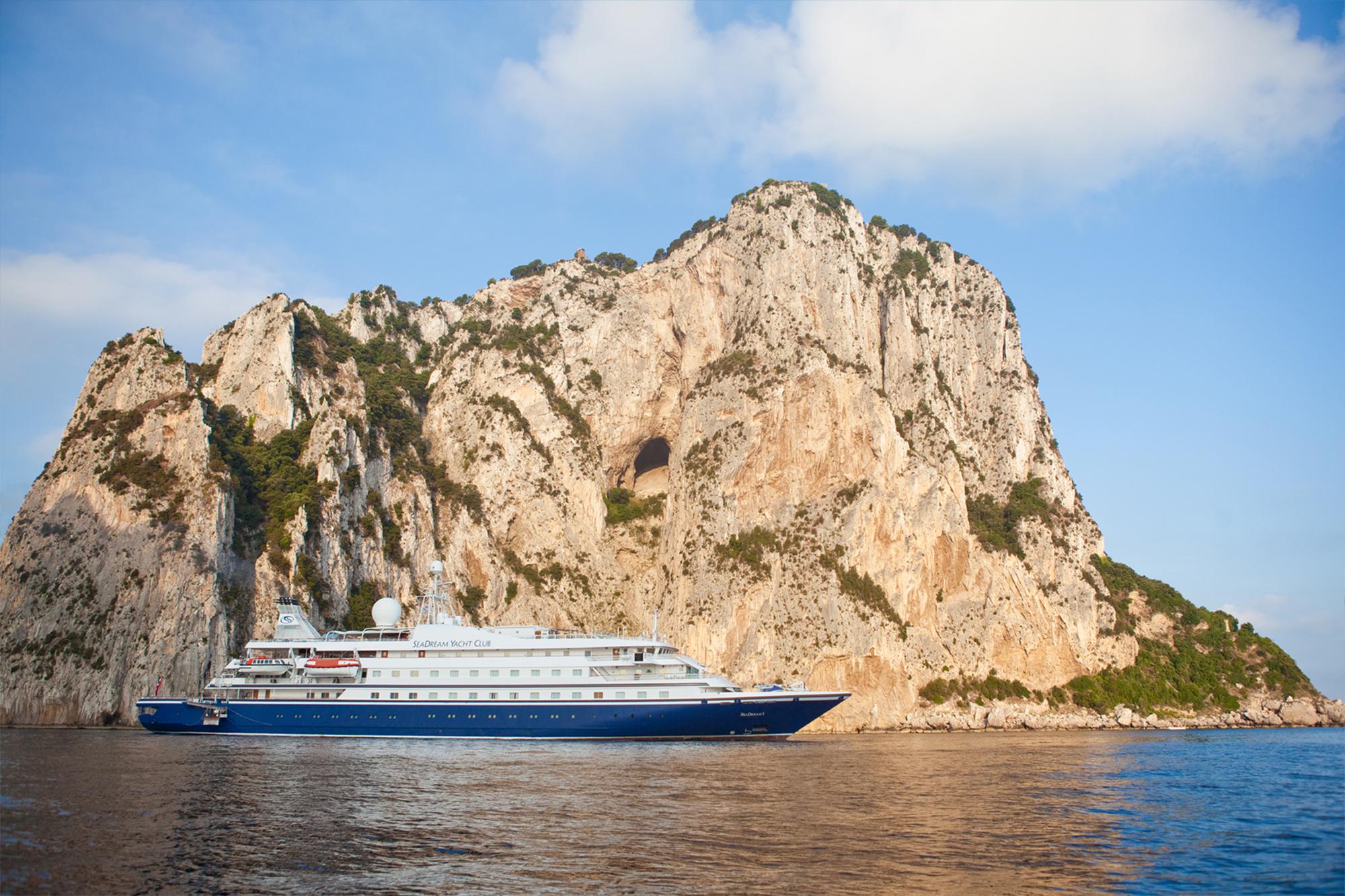 A large multi-deck cruise ship anchors in the azure waters off Capri, Italy, positioned against the dramatic limestone cliffs and natural rock formations of the Faraglioni. The vessel's white superstructure and blue hull contrast sharply with the rugged, tan-colored geological formations dotted with Mediterranean vegetation.