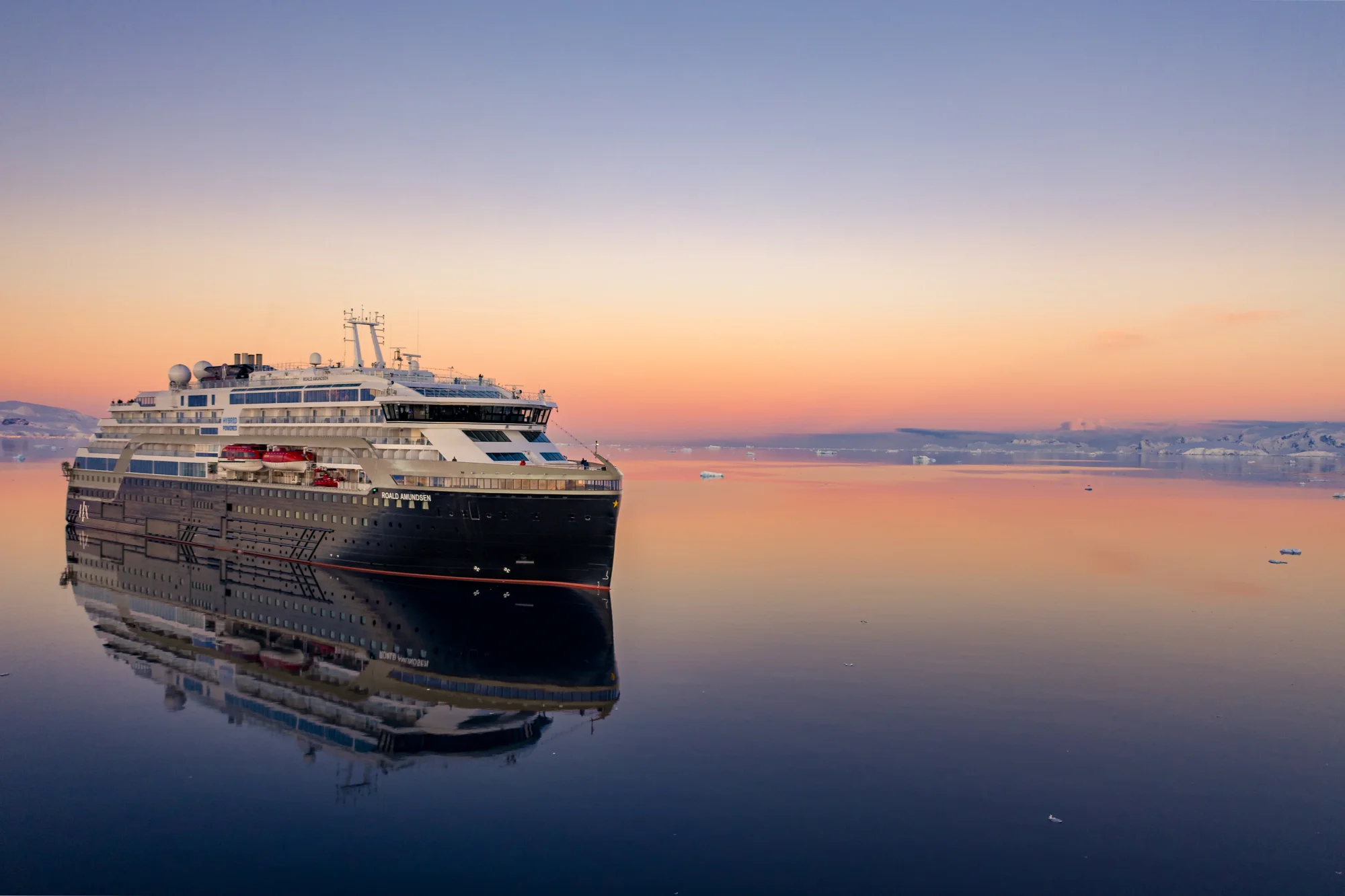 A large modern cruise ship is anchored in calm, mirror-like waters during a stunning golden sunrise, with its distinctive black hull and white upper decks perfectly reflected in the glassy sea. The peaceful morning scene captures the ship's impressive multi-deck architecture against a softly colored sky transitioning from warm orange and pink to cool blue.