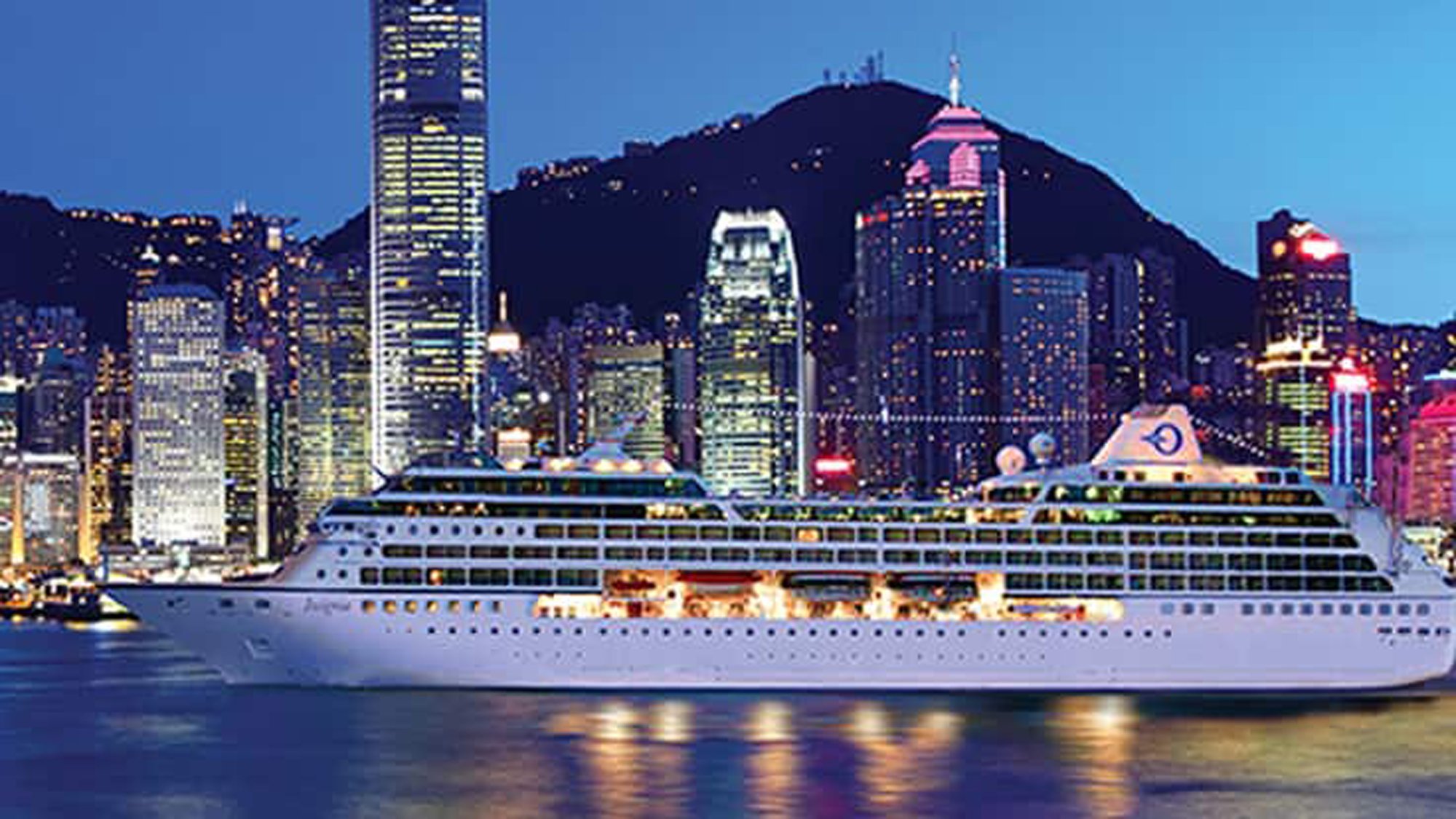A white luxury cruise ship is moored in Hong Kong's Victoria Harbour during twilight, with the illuminated skyline of skyscrapers and mountains rising dramatically in the background. The scene captures the vibrant cityscape with its distinctive architecture and harbor waters reflecting the ship's lights.