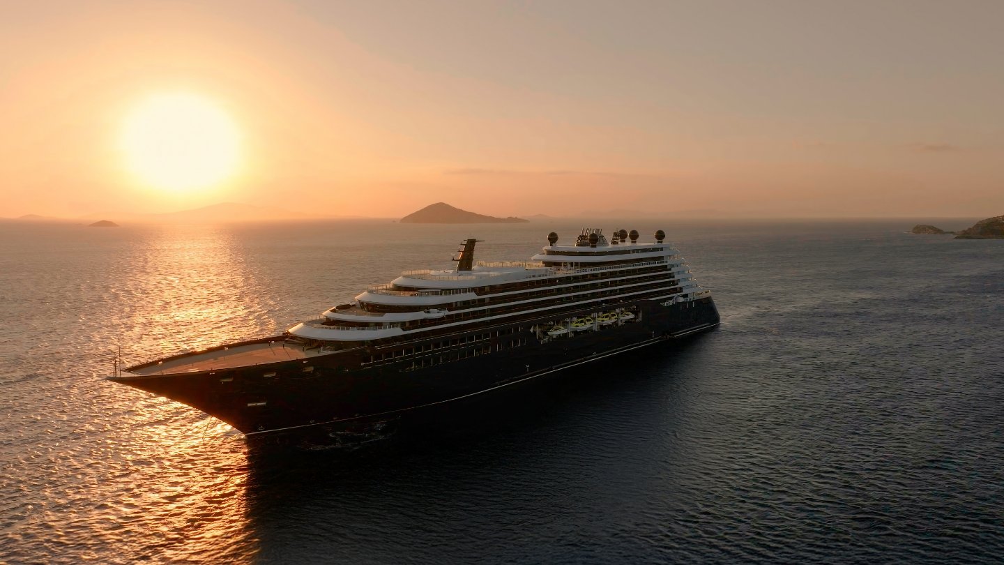 A modern large cruise ship anchored in calm Mediterranean waters at sunrise, with warm golden light reflecting off the sea and distant islands visible in the hazy background. The vessel features multiple white deck levels contrasting against a dark hull, photographed during the golden hour with a serene, peaceful atmosphere.