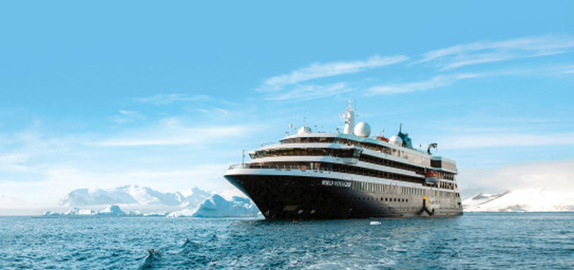 A large expedition cruise ship navigates through icy Antarctic waters with snow-capped mountains and glaciers visible in the background. The vessel displays multiple decks and communication equipment against a clear blue sky, showcasing the scale and sophistication of modern polar cruising.
