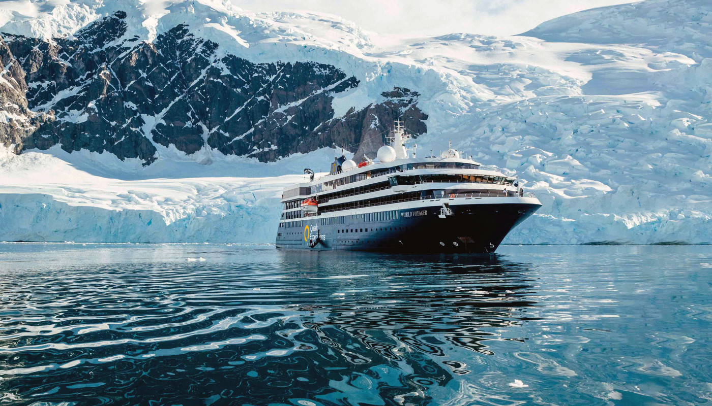 A large expedition cruise ship navigates through pristine Antarctic waters surrounded by massive icebergs and snow-capped mountain peaks. The vessel is positioned against a dramatic backdrop of glaciers and jagged rock formations, showcasing the ultimate polar expedition experience.