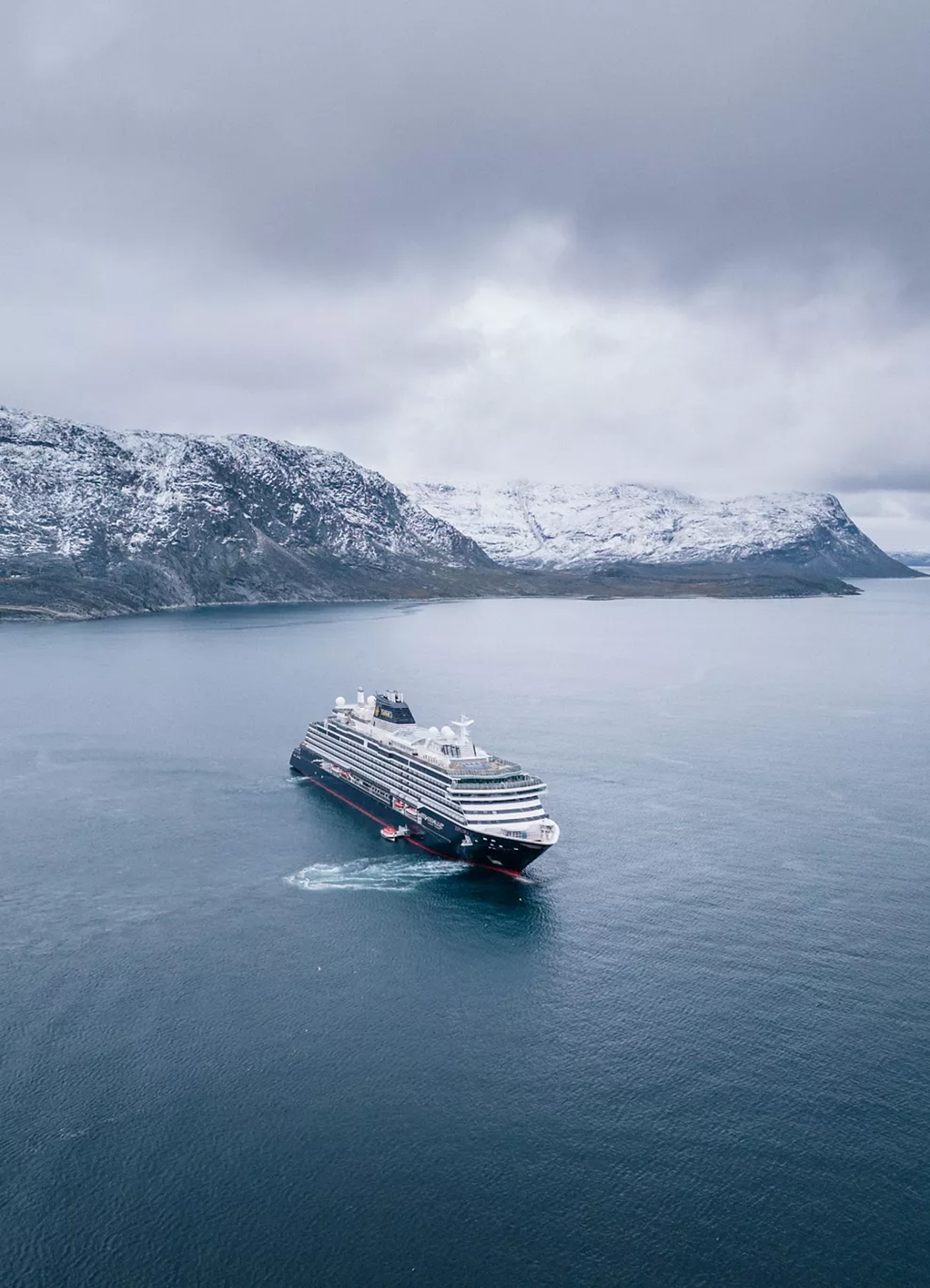 Luxury Cruise Ship in Arctic Waters A modern expedition cruise ship navigates through pristine Arctic waters with snow-capped mountains rising dramatically in the background. The vessel is surrounded by calm seas under overcast skies, showcasing a remote Nordic landscape typical of Arctic cruising destinations.