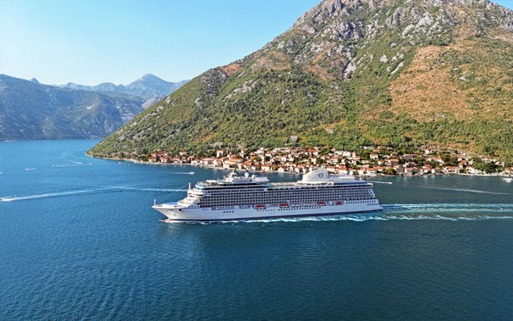 A modern large cruise ship navigates through a stunning Mediterranean fjord landscape, with a picturesque waterfront village nestled against steep, forested mountain slopes. The crystalline blue waters and dramatic alpine scenery create an iconic Mediterranean cruising destination.