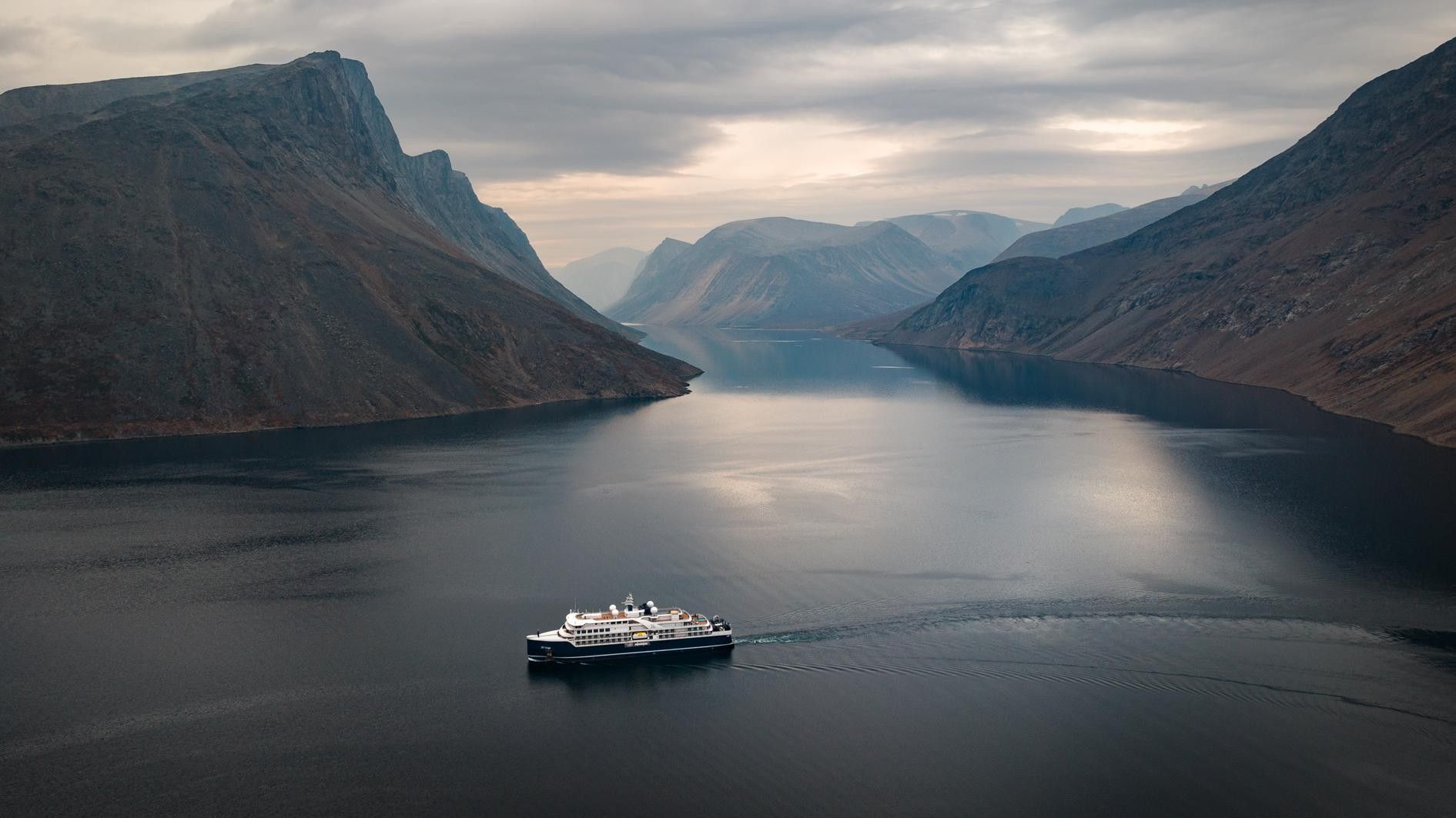A modern expedition cruise ship navigates through a dramatic Norwegian fjord surrounded by steep, snow-capped mountains and calm glacial waters. The vessel is dwarfed by the towering peaks that frame this pristine Arctic landscape, showcasing the scale and beauty of Scandinavian fjord exploration.