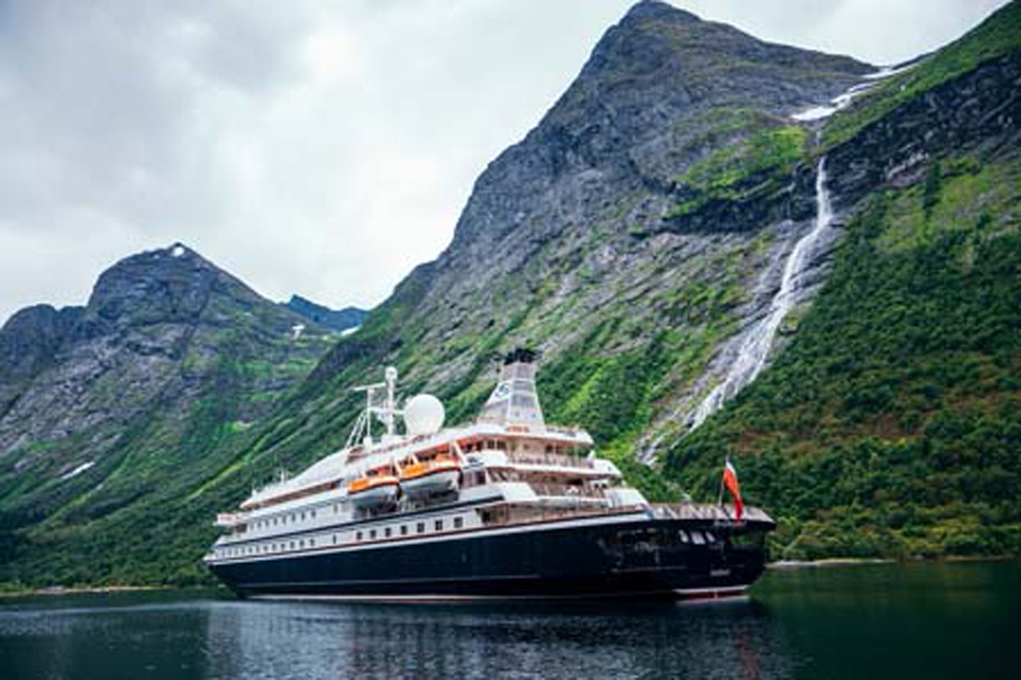 A large expedition cruise ship anchors in a dramatic Norwegian fjord, surrounded by steep mountainsides covered in green vegetation and cascading waterfalls. The vessel features a distinctive white superstructure with dark hull, positioned against a backdrop of towering peaks and pristine alpine scenery.