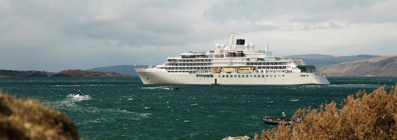 A white luxury cruise ship anchored in a turquoise ocean with rocky coastline and mountains in the background. Small boats and watercraft are visible near the vessel.